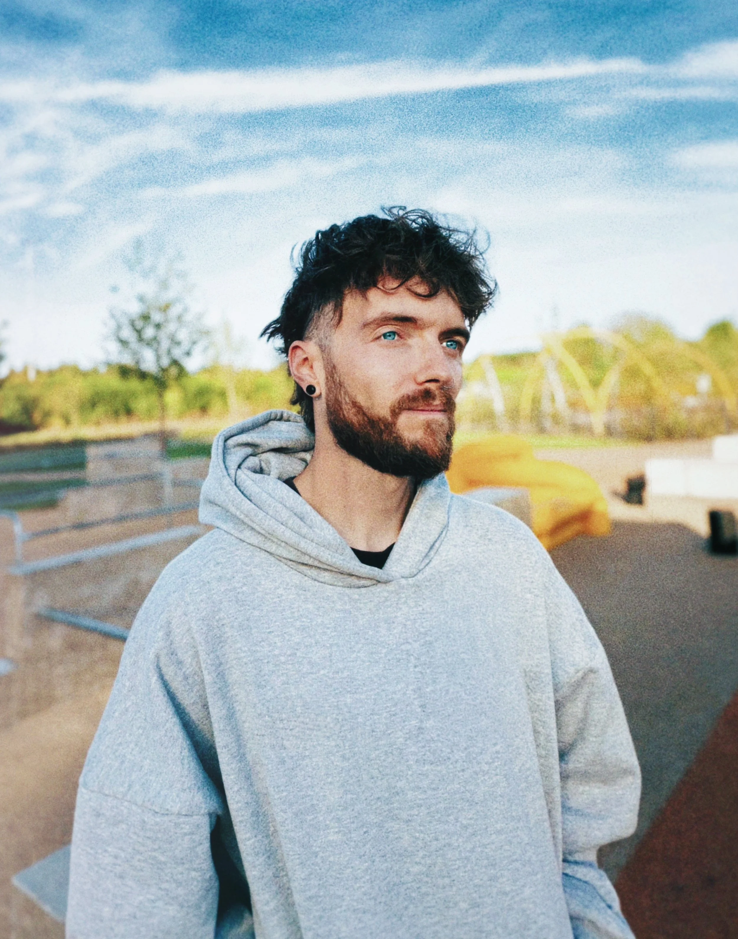 A young man with curly hair and a beard, wearing a grey T-shirt, standing outdoors near a brick building and parking area under an overhang.