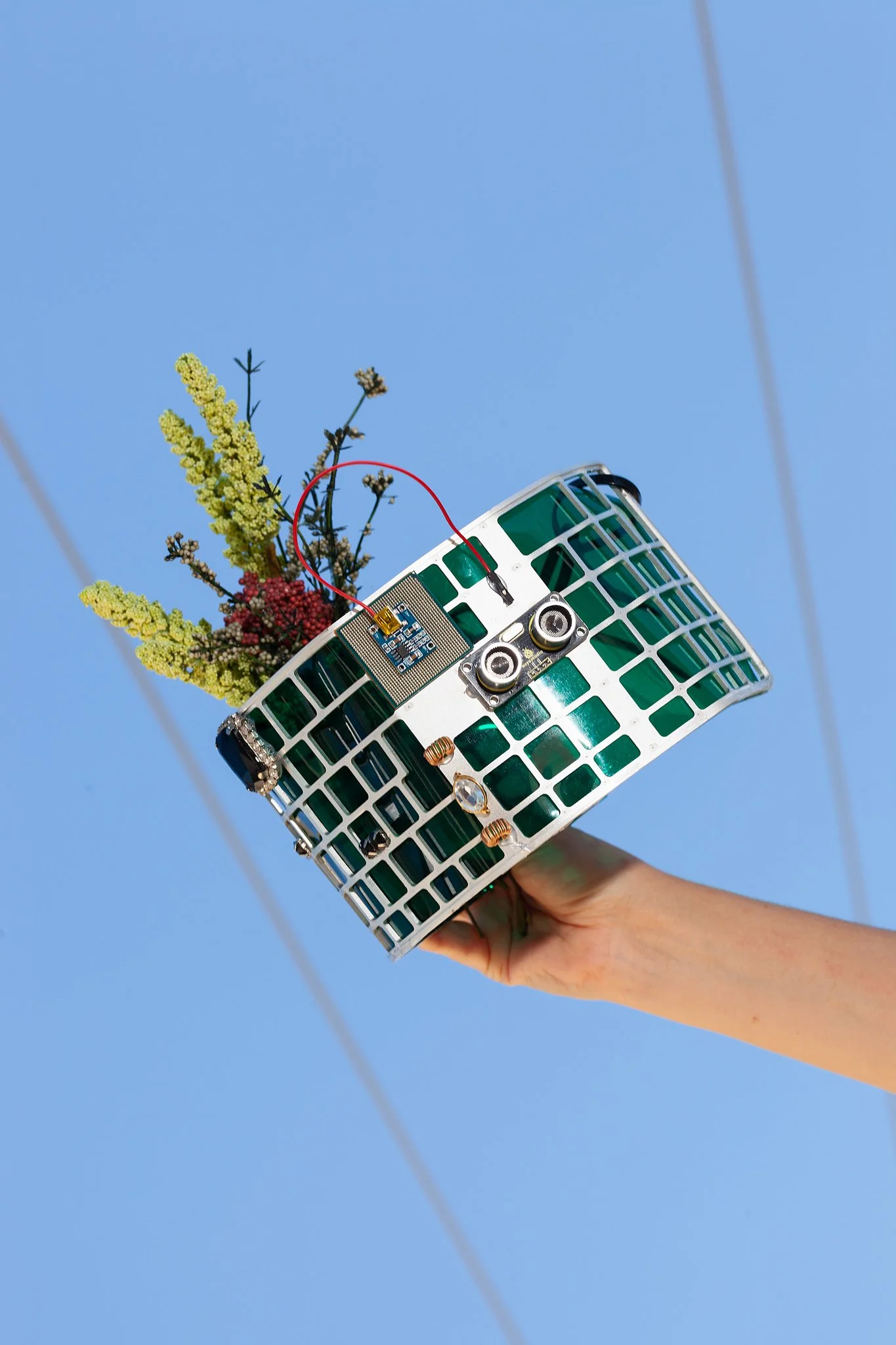 Hand holding a basket with electronic components and flowers against a blue sky.