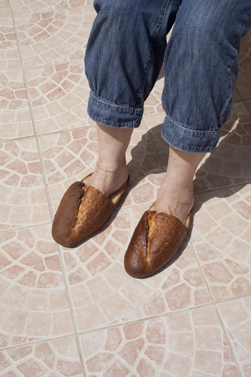 Person wearing oversized bread loaf slippers and rolled-up blue jeans standing on tiled floor.