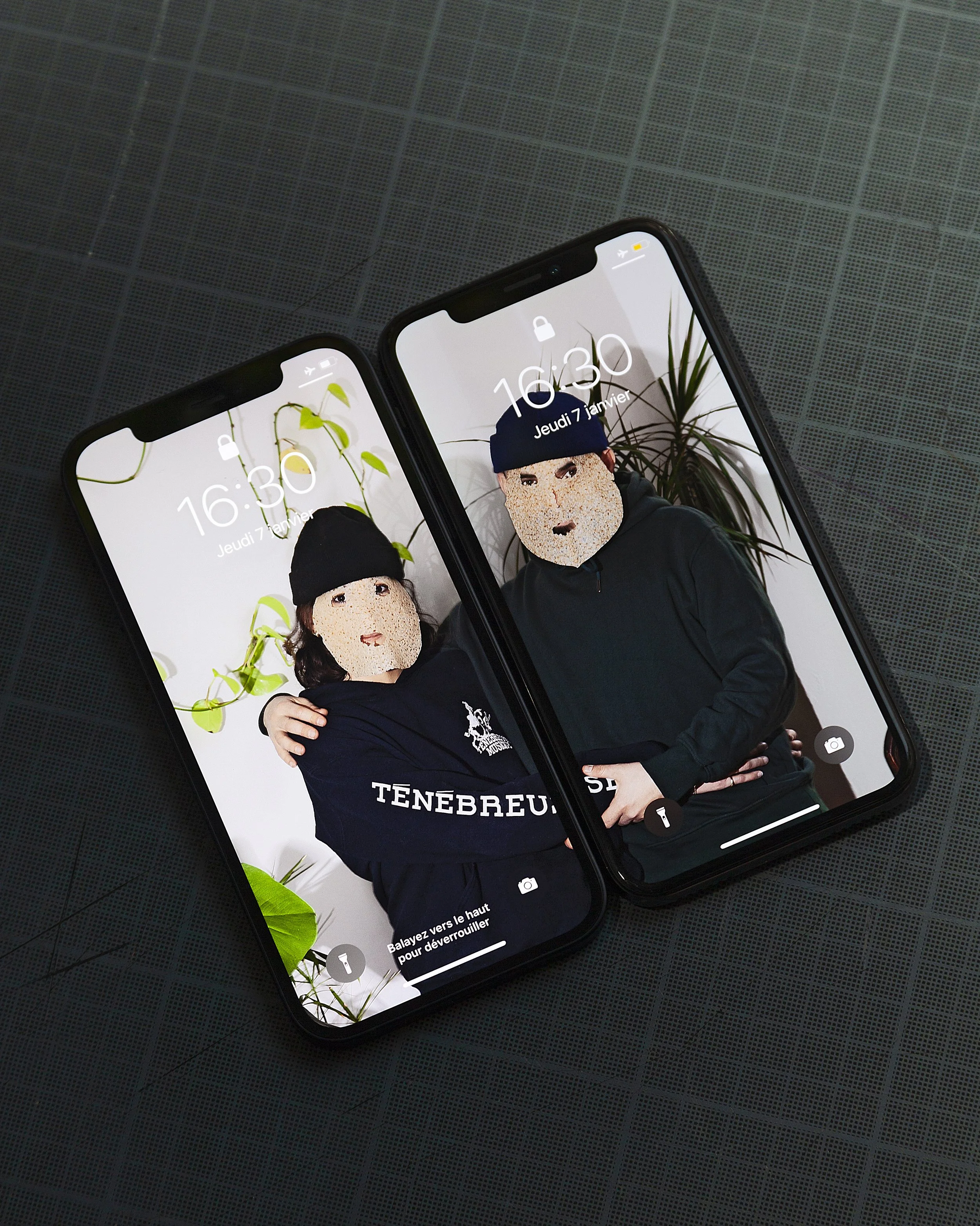 Two smartphones displaying locked screens with photos of two people wearing black beanies and dark clothing, standing in front of a white wall with green plants.
