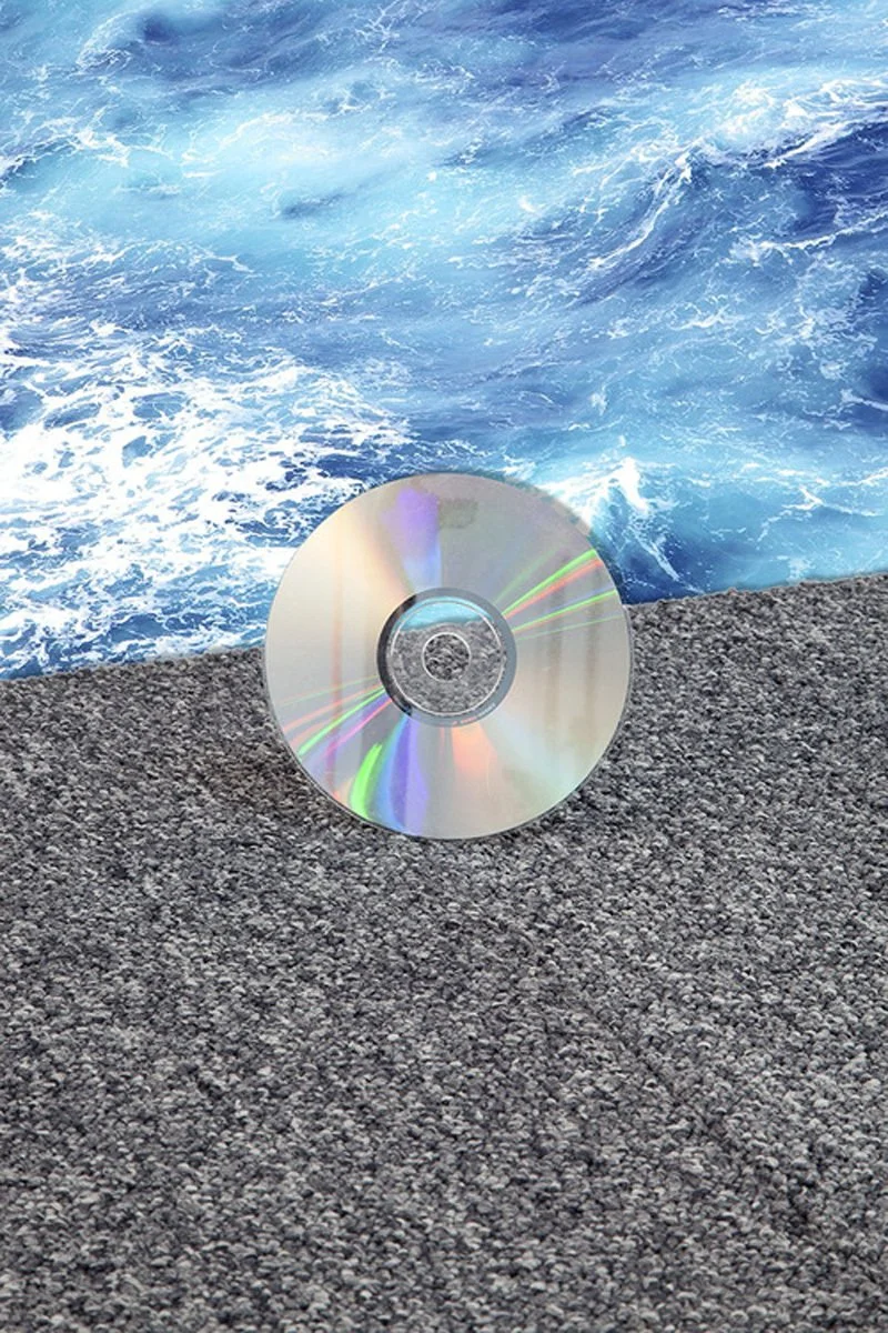 A compact disc resting on a dark gray, textured surface with a background of ocean water with waves.