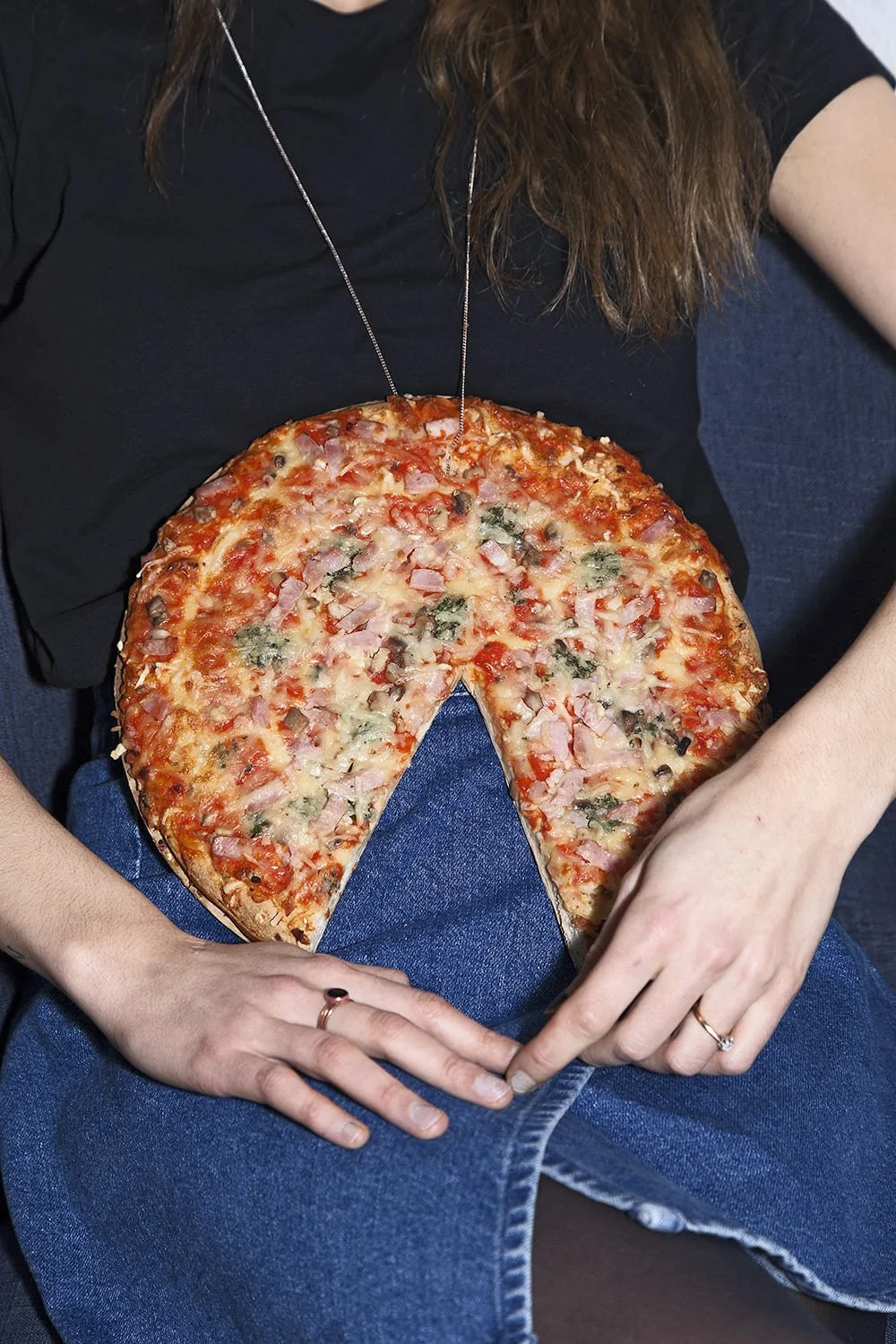 Person wearing a black shirt and jeans sitting on a dark sofa, holding a large pizza with one slice removed.