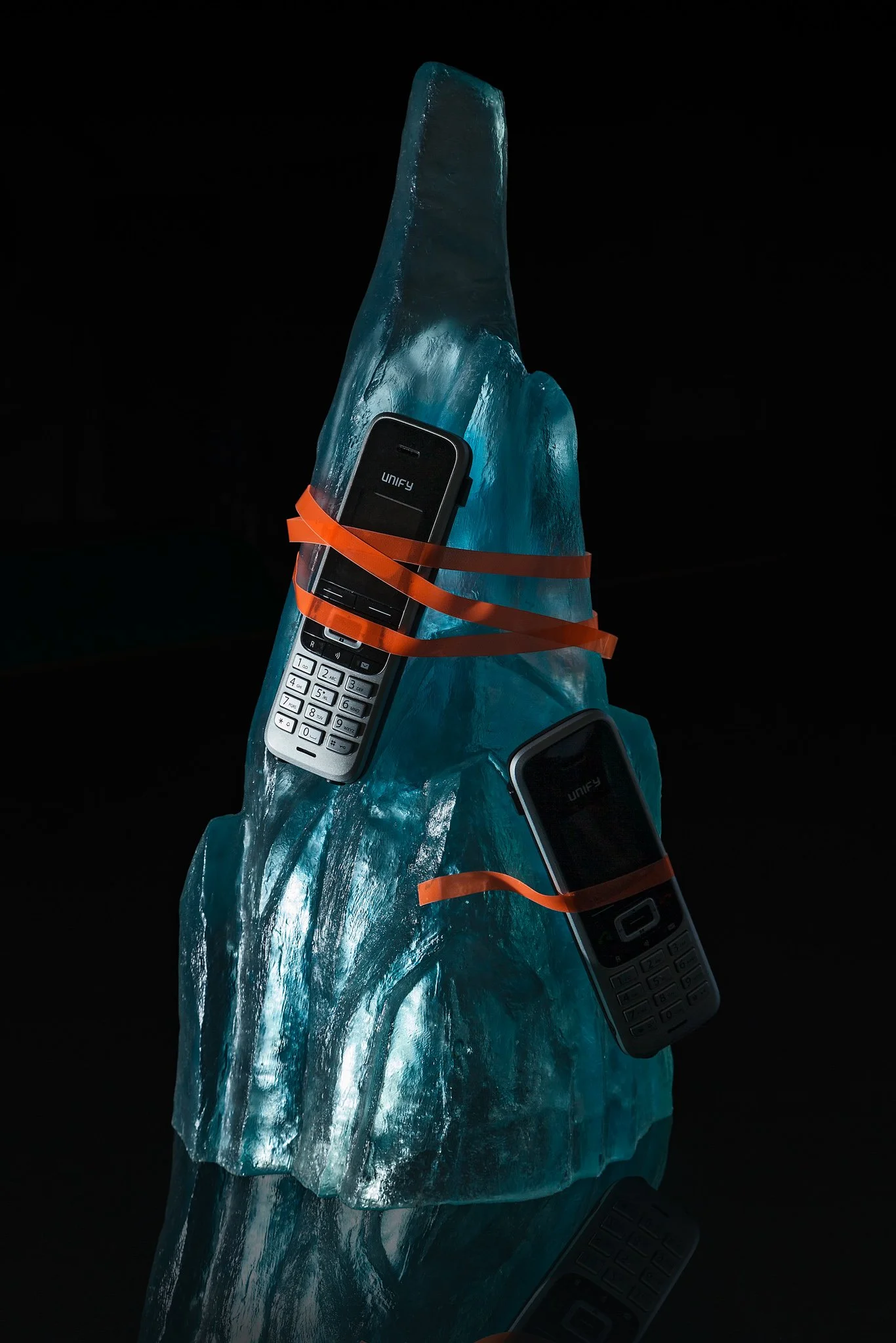 Two old mobile phones secured with orange rubber bands to a large blue-green ice sculpture with a reflective surface.
