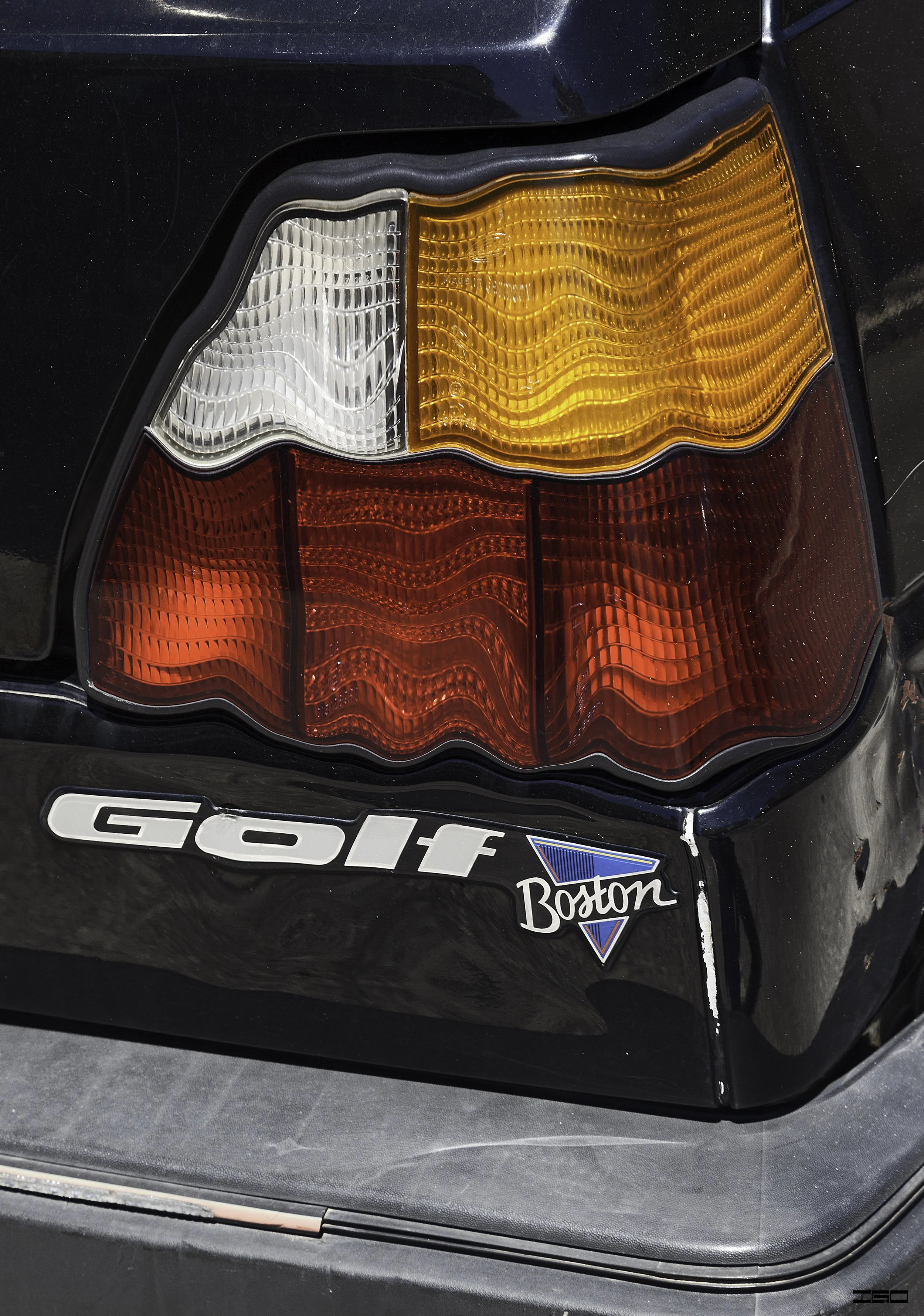 Close-up of the rear taillight of a black car with stickers that say 'GOLF' and 'Boston'.