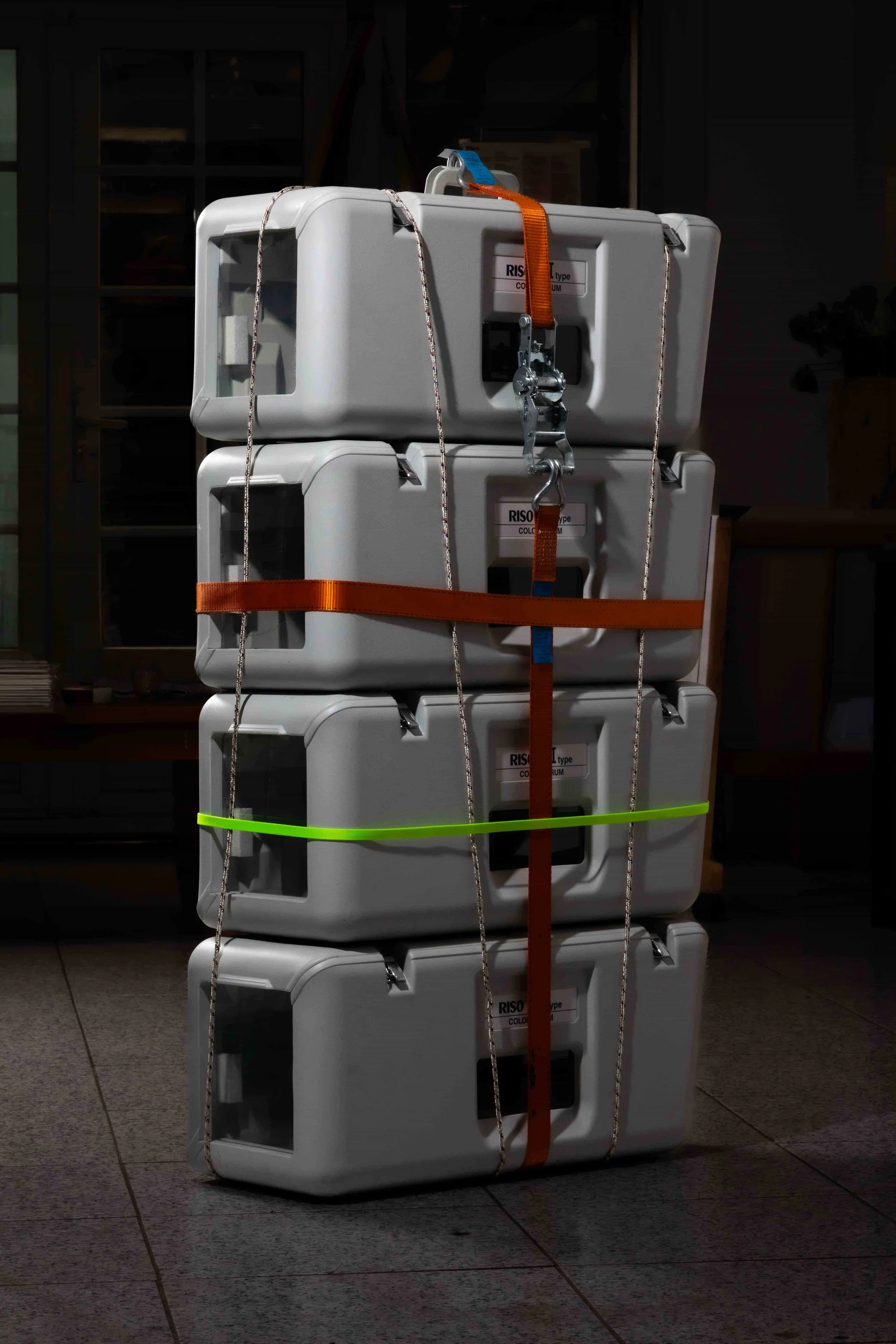 A stack of five large gray plastic containers secured with colorful straps and metal hooks.