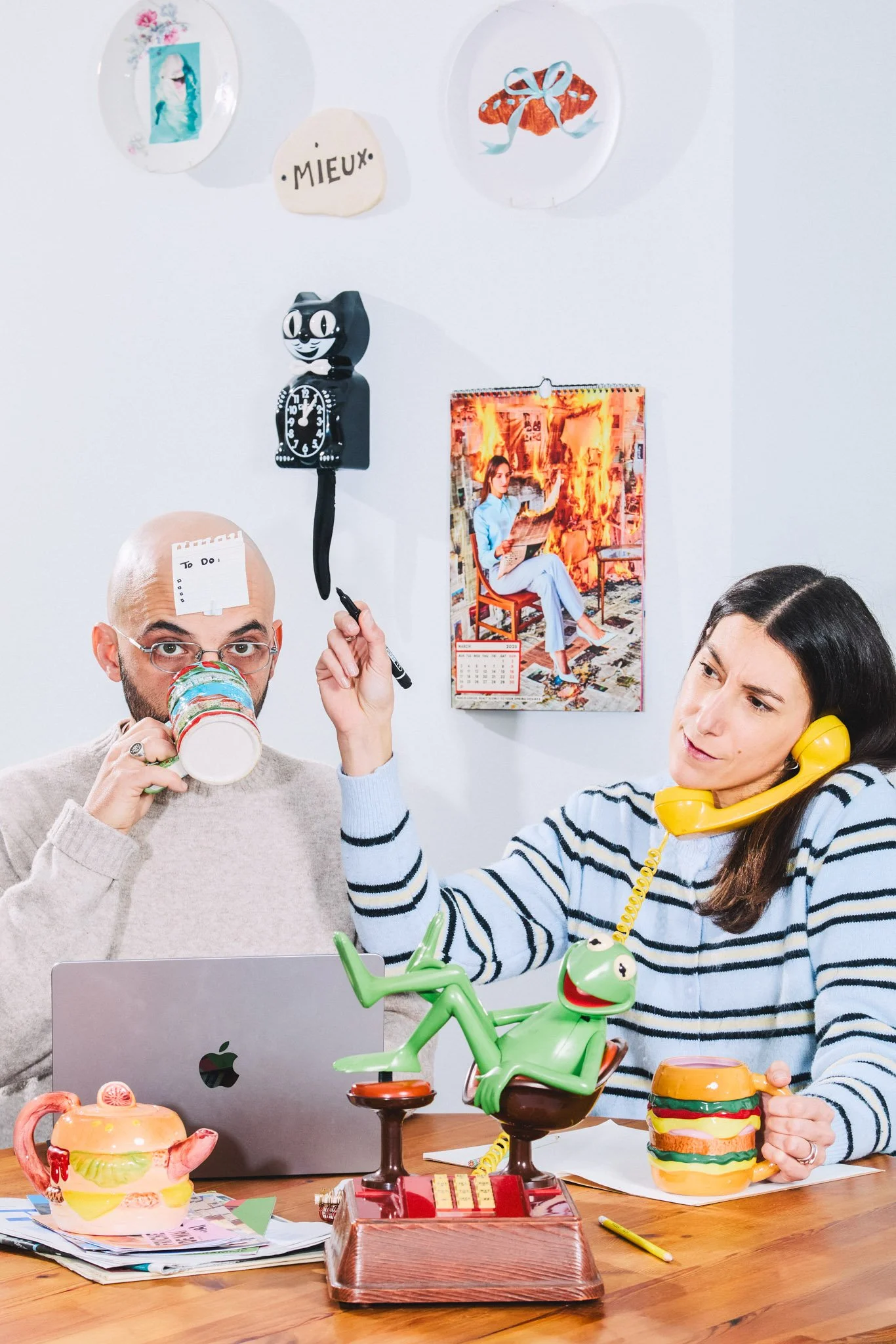 Two people sitting at a table cluttered with colorful ceramics, a laptop, and papers, with a decorative clock and calendar on the wall behind them. One person is drinking from a colorful mug, and the other is talking on a yellow rotary phone. There i