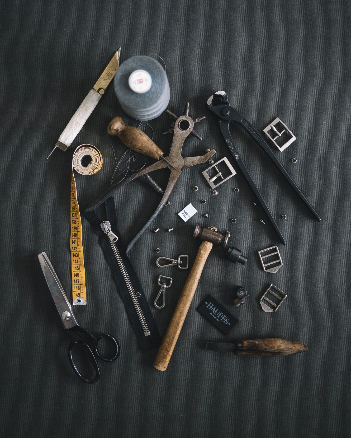 Assorted tools including scissors, a hammer, nails, zippers, a measuring tape, screws, and other small hardware on a dark surface.