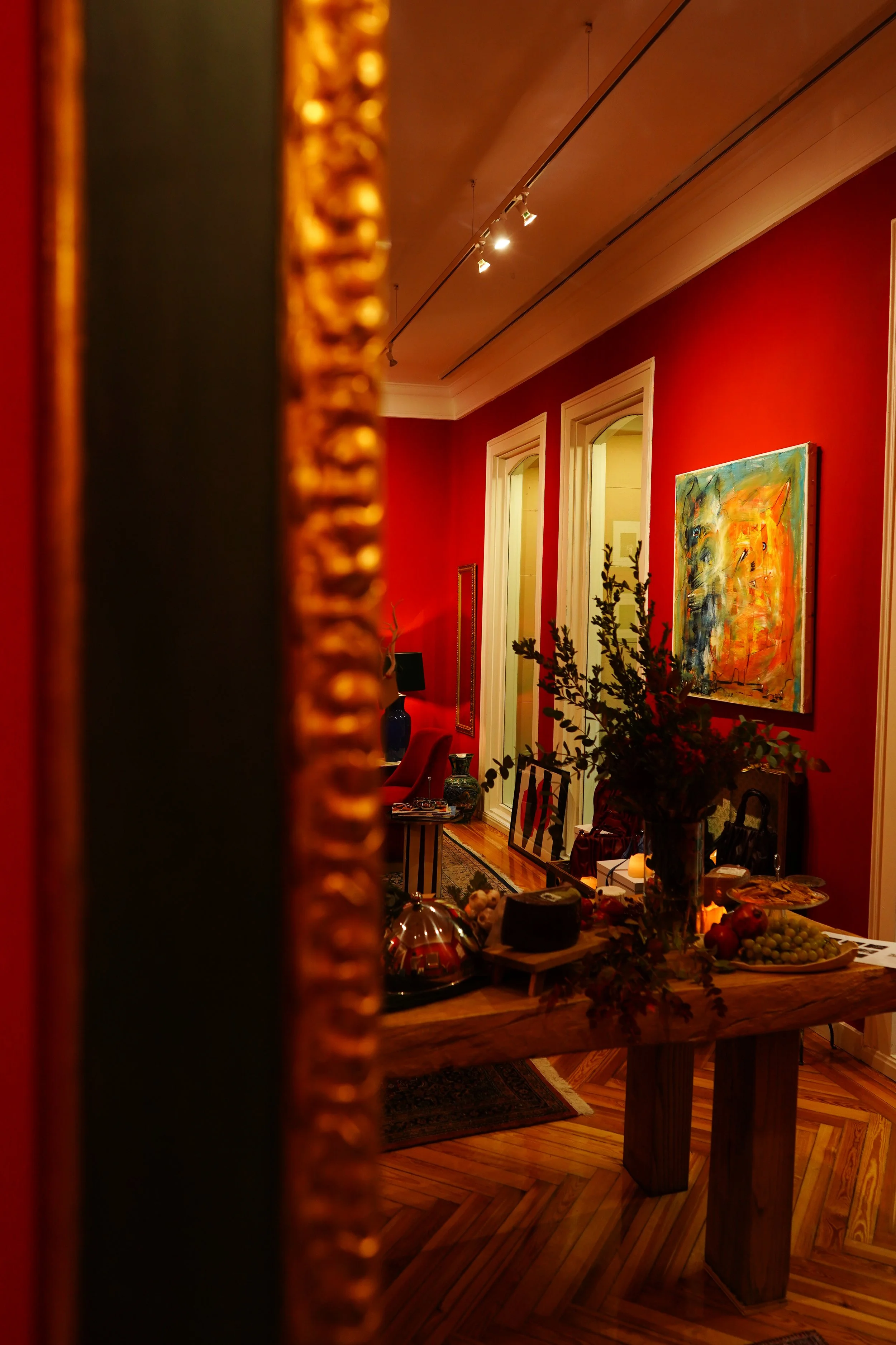 The image shows a room with red walls and a wooden ceiling, viewed partially through a mirror with a decorative gold frame. The room has a table with various items, including a vase shaped like a person, what appears to be food, and other decorative objects. There is also a painting on the wall depicting an abstract colorful scene, and some greenery in the foreground.