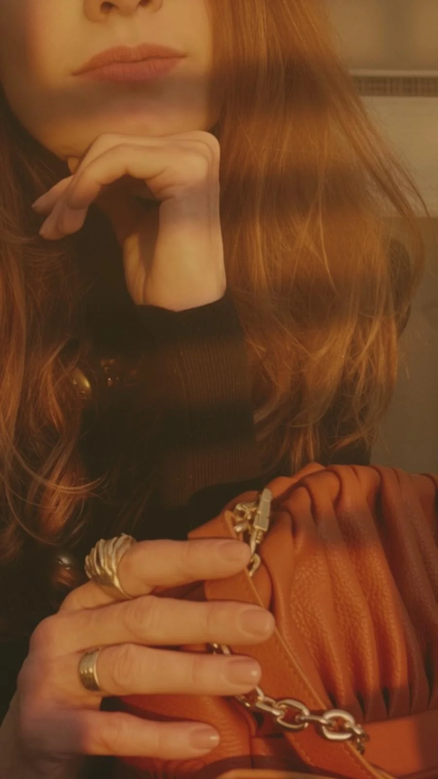 Close-up of a woman's face and hands. She rests her chin on her hand, which has a silver ring, and holds a tan leather bag with a silver chain strap. She has long reddish hair, a black top, and is in a dimly lit setting.