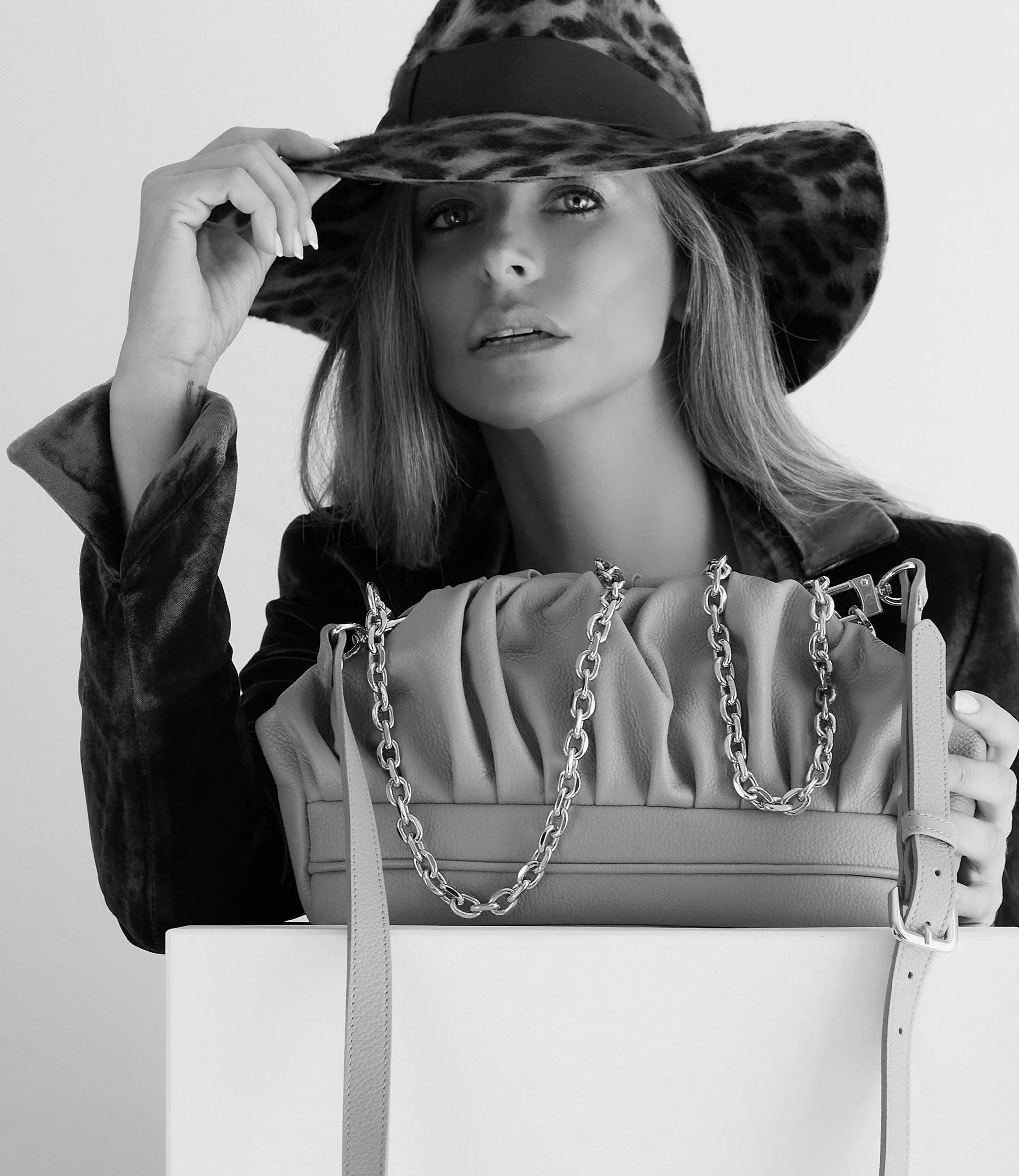 A woman wearing a wide-brimmed leopard print hat, holding the brim, with a chain-strap bag in front of her, posing behind a box in a black-and-white fashion photograph.