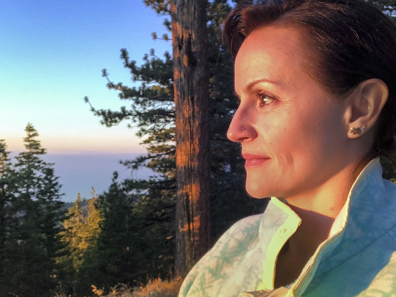 A woman with short dark hair, Vision Quest Facilitator Tina, is looking towards the sunrise in a forested area.