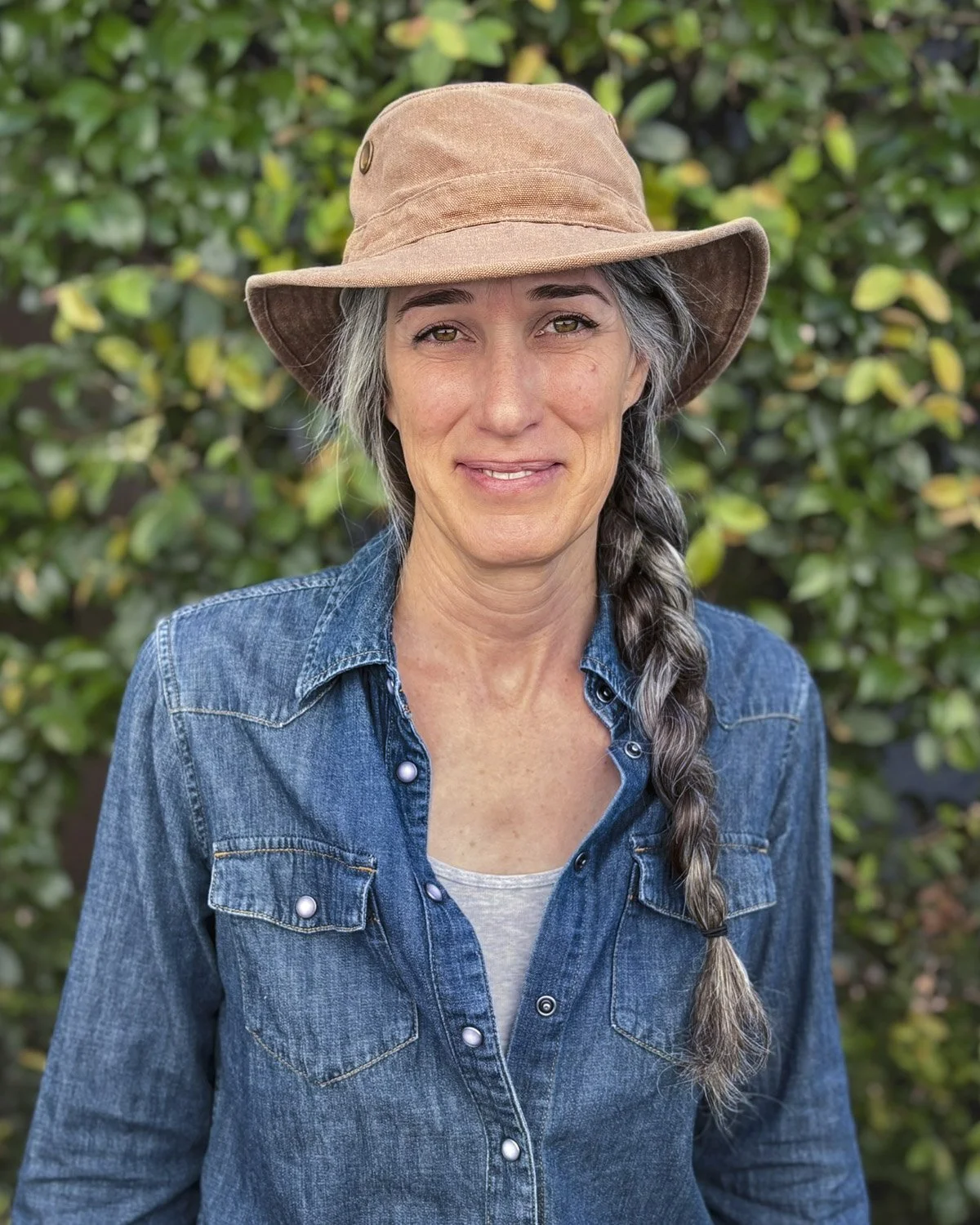 Educator and Vision Quest Facilitator, Tanya, wearing a wide-brimmed brown hat, denim shirt, and white top, standing outdoors in front of green foliage.