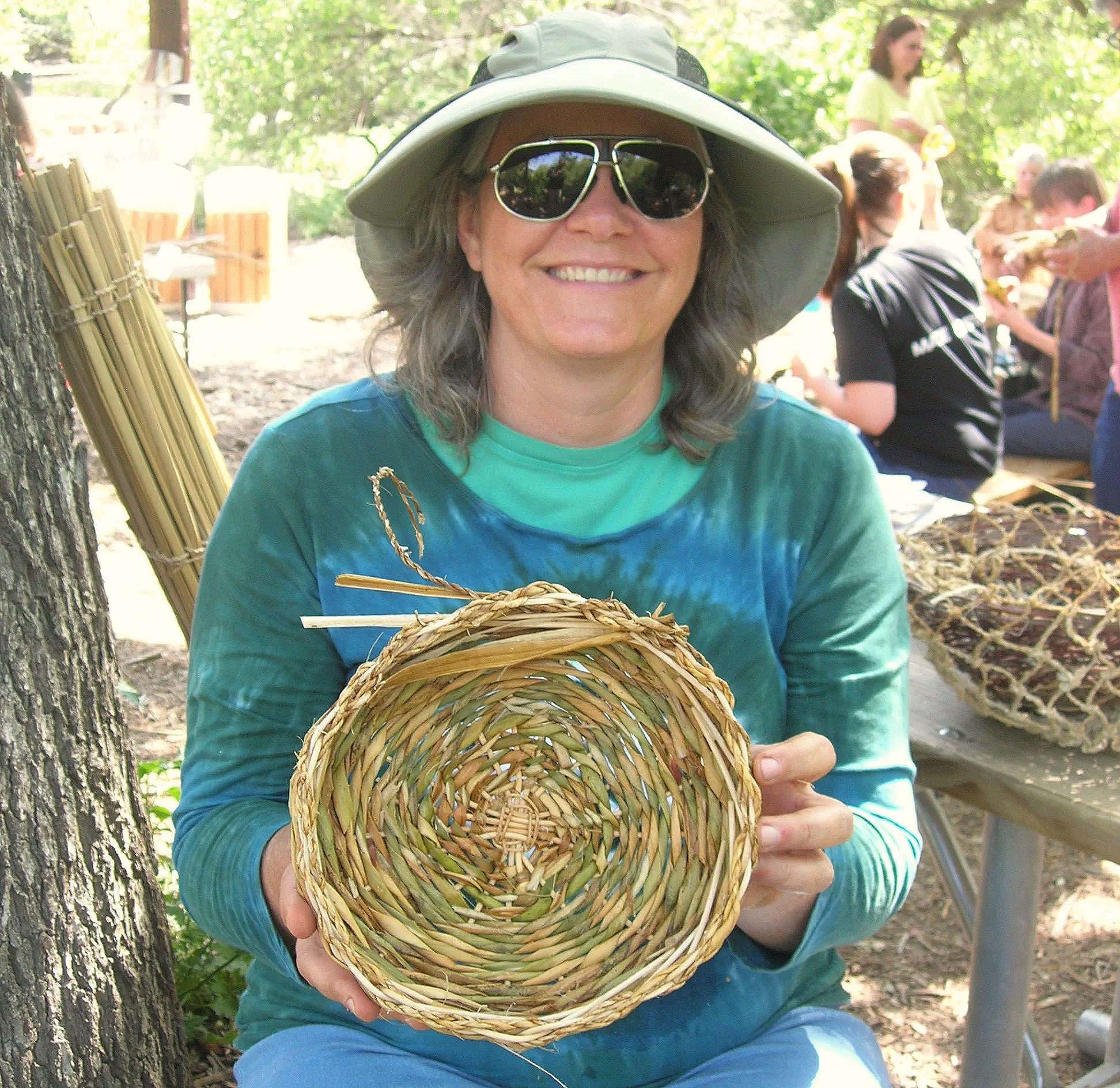 Workshop participant smiling wearing sunglasses and a sunhat, holding a hand woven basket outdoors with other students in the background.