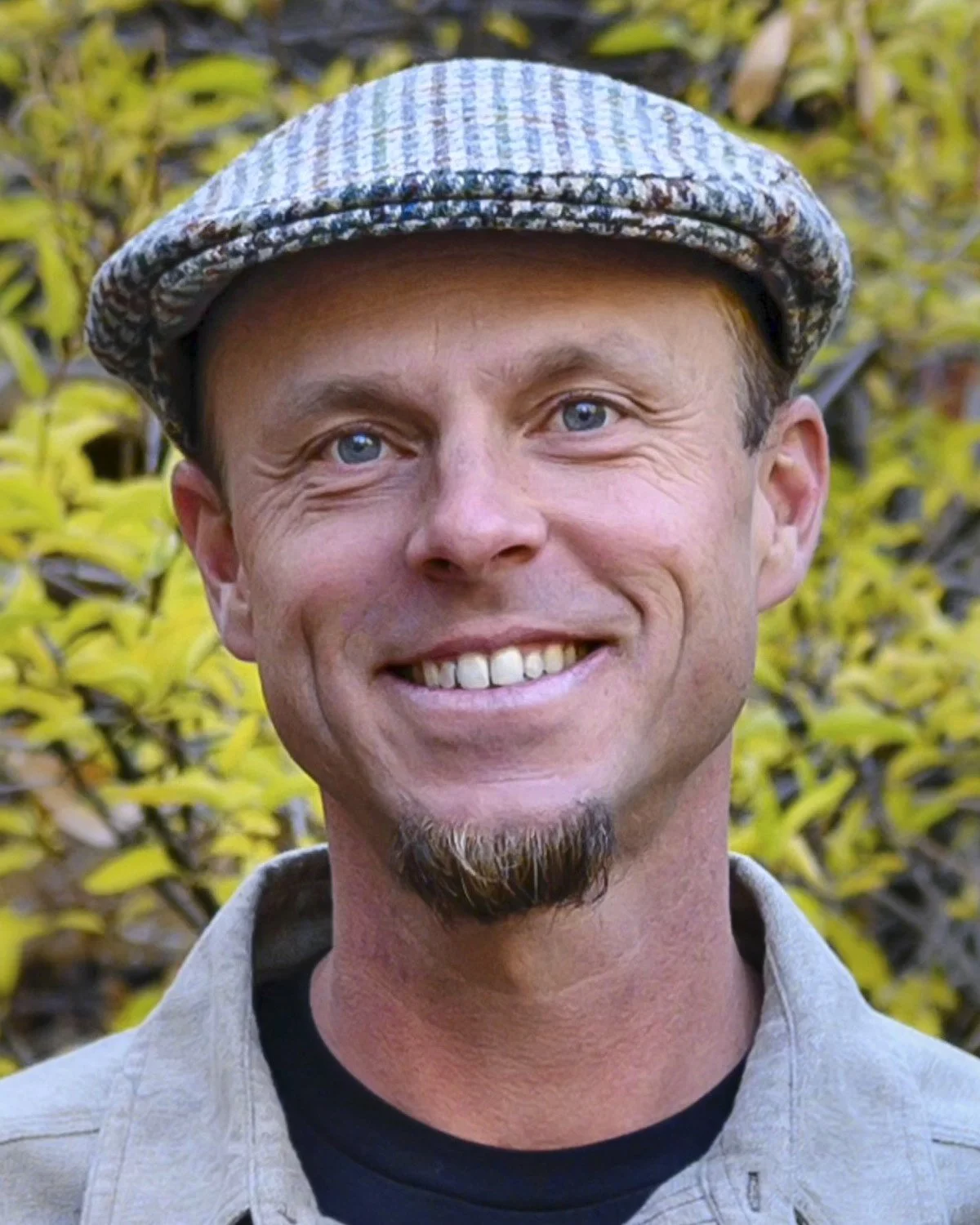 A man with blue eyes and a goatee, Daniel - educator and Vision Quest Facilitator, wearing a flat cap and smiling in front of a background of yellow-green foliage.