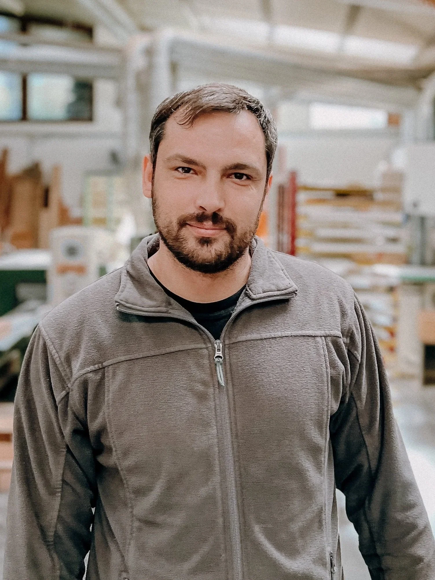 Mann in einer Werkstatt trägt eine graue Jacke. Im Hintergrund sind Holzarbeiten und Maschinen sichtbar.