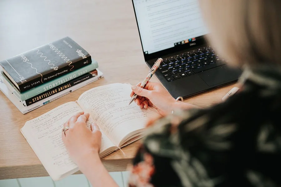 A woman holding a pen and writing in her notebook, next to her laptop.