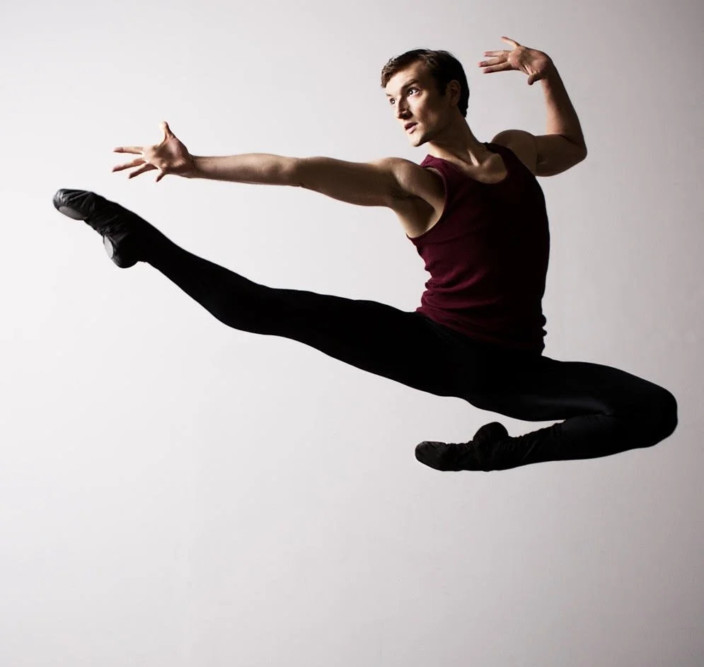 A male dancer performing a high leap, with one leg extended forward and the other bent beneath him, arms outstretched, wearing a maroon sleeveless top and black pants against a plain light background.