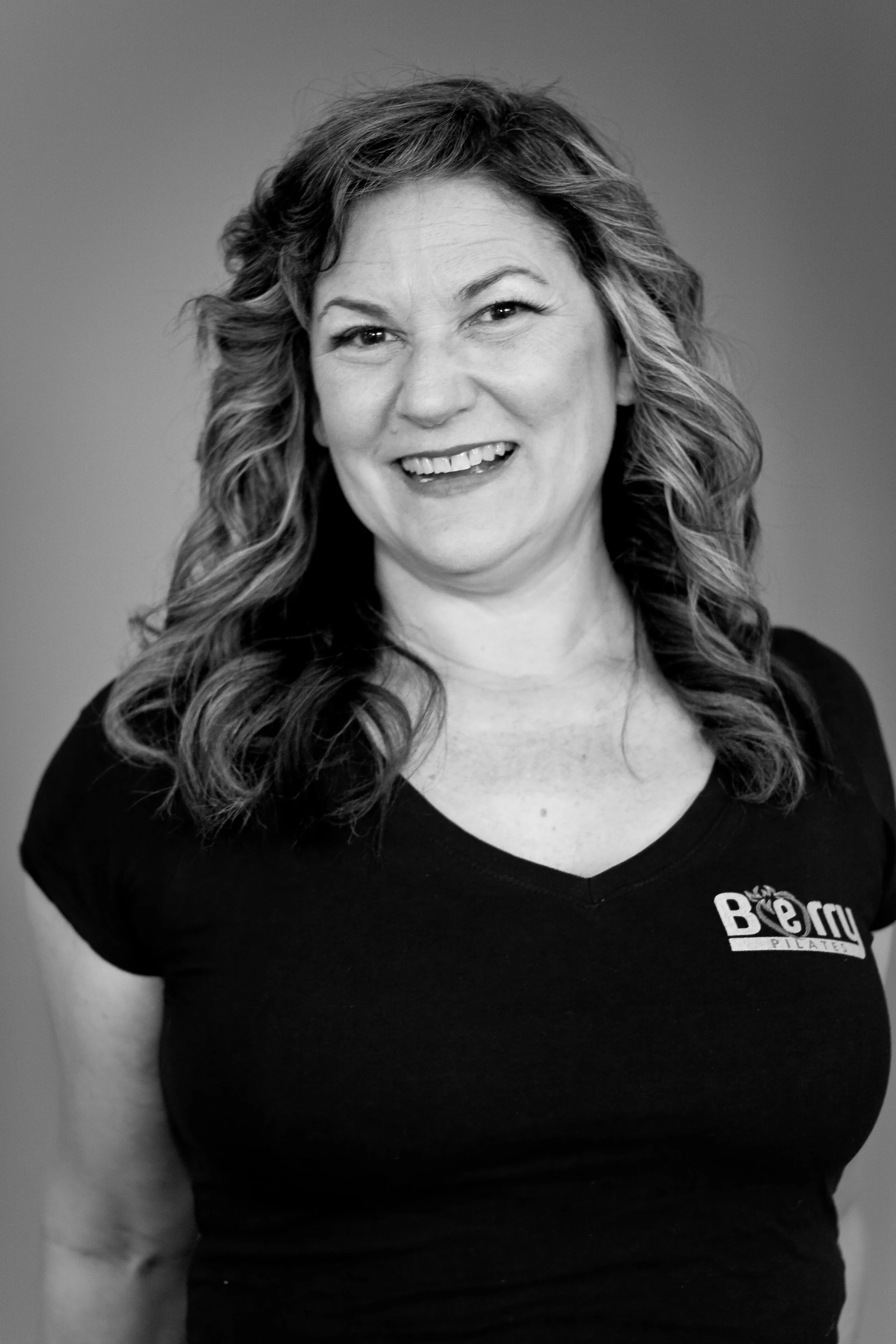 A smiling woman with wavy hair wearing a black T-shirt with a logo that says 'B e r y' in a studio setting.