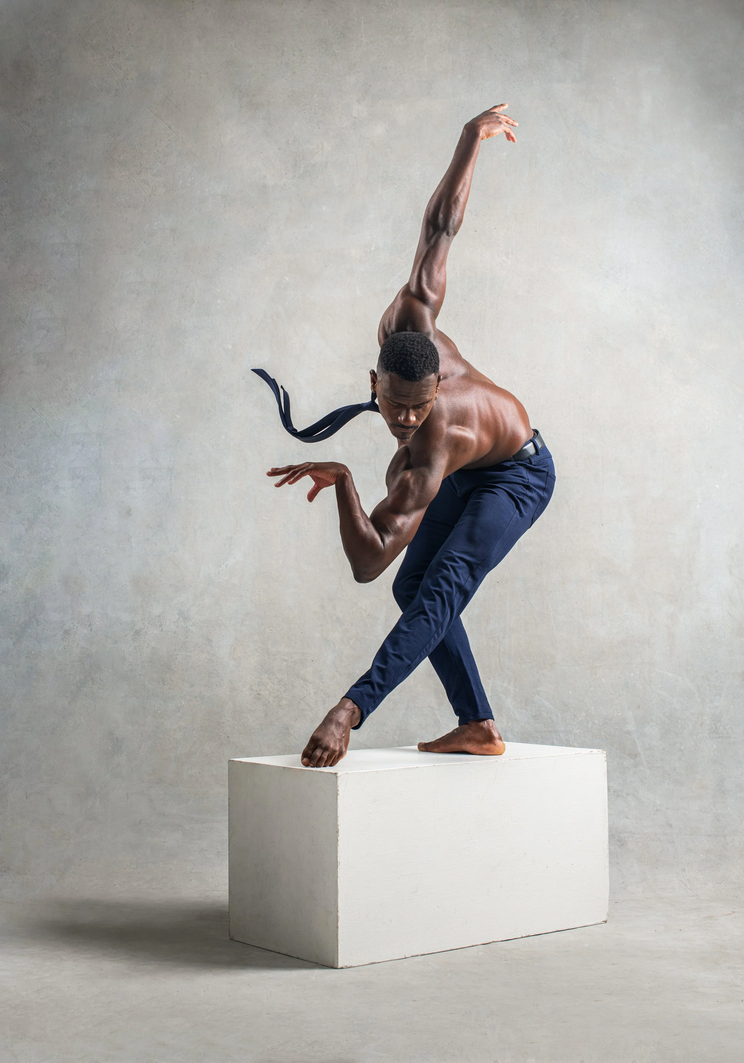 A shirtless man with short hair and dark pants is balancing on a white rectangular block, with one foot on the block and the other foot and one hand extended downward, and the other arm raised upward.