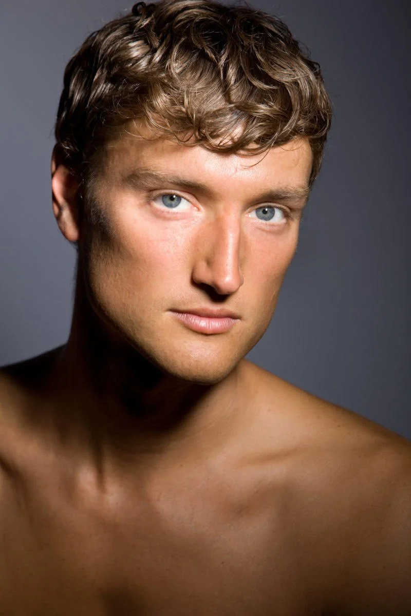 Portrait of a young man with short, wavy brown hair and blue eyes, looking at the camera with a slight smile, against a plain gray background.