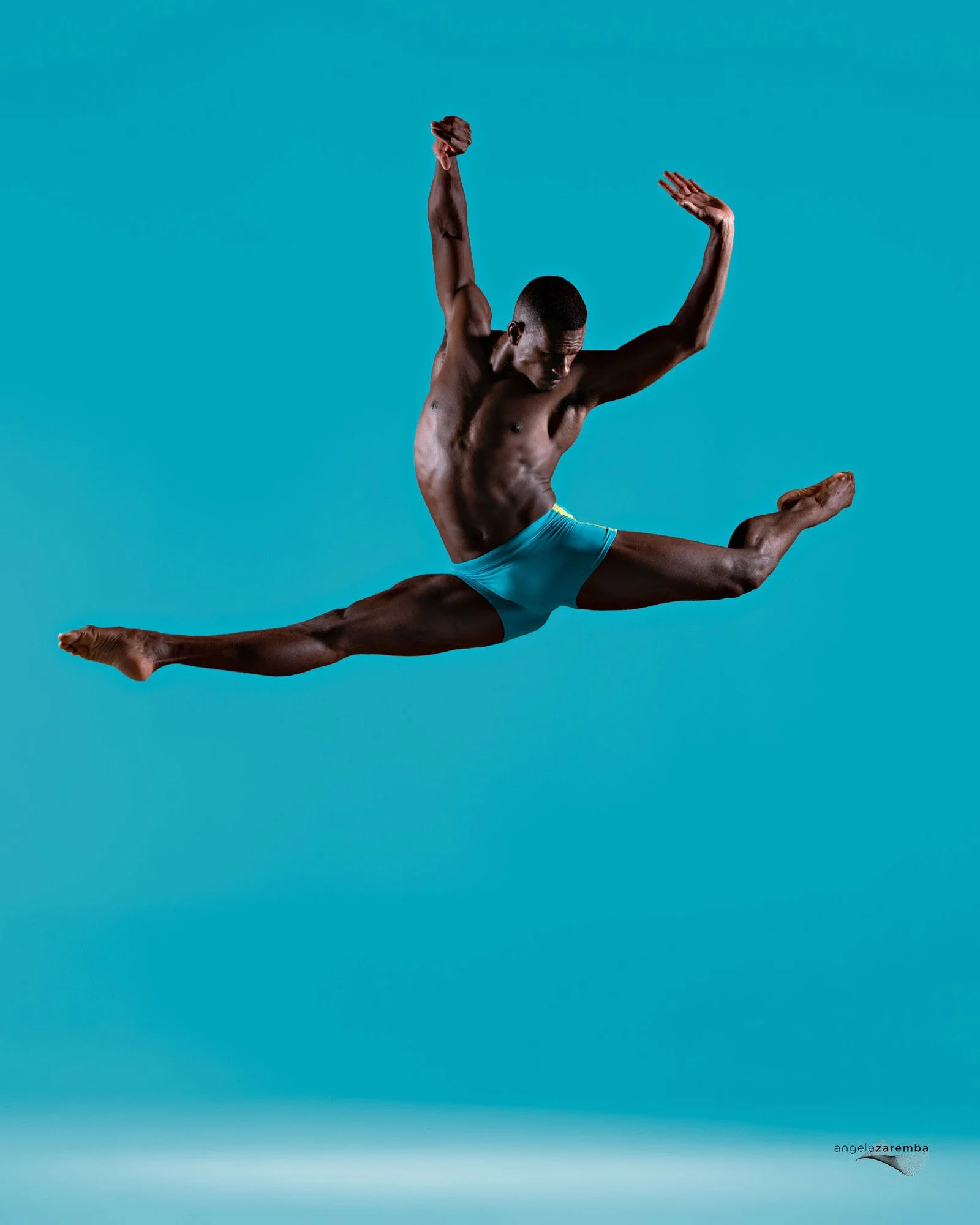A male gymnast in mid-air performing a split leap with arms raised and legs extended against a solid light blue background.