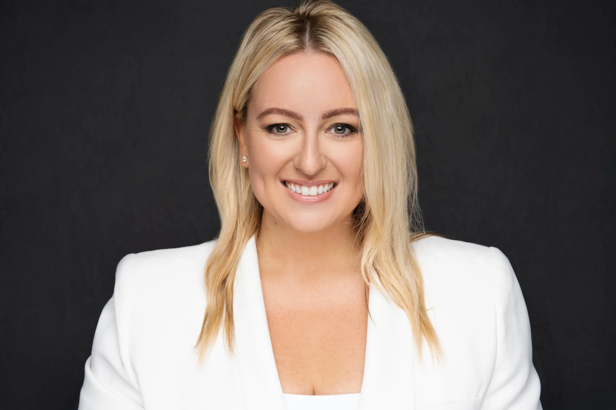 Professional woman with blonde hair, white blazer, and a friendly smile, posing against a dark background.