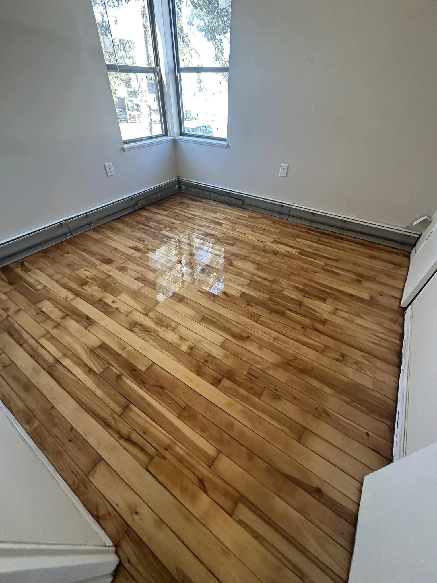 Empty room with polished wooden floor, white walls, and large windows showing street view outside.