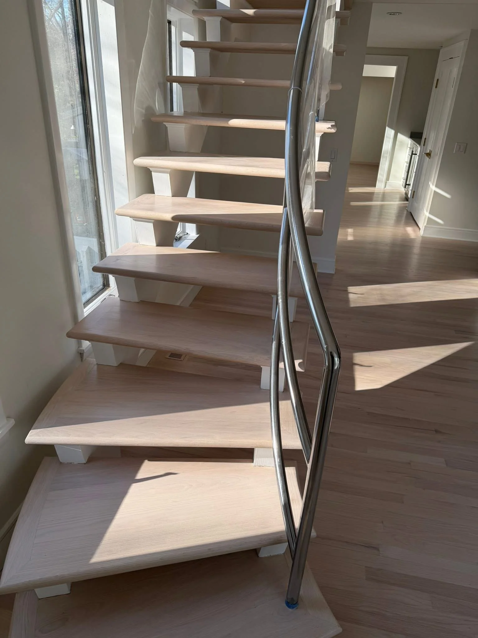 Interior view of a wooden staircase next to large windows, with sunlight casting shadows on the stairs and floor, leading into a bright room with white walls and open doorways.