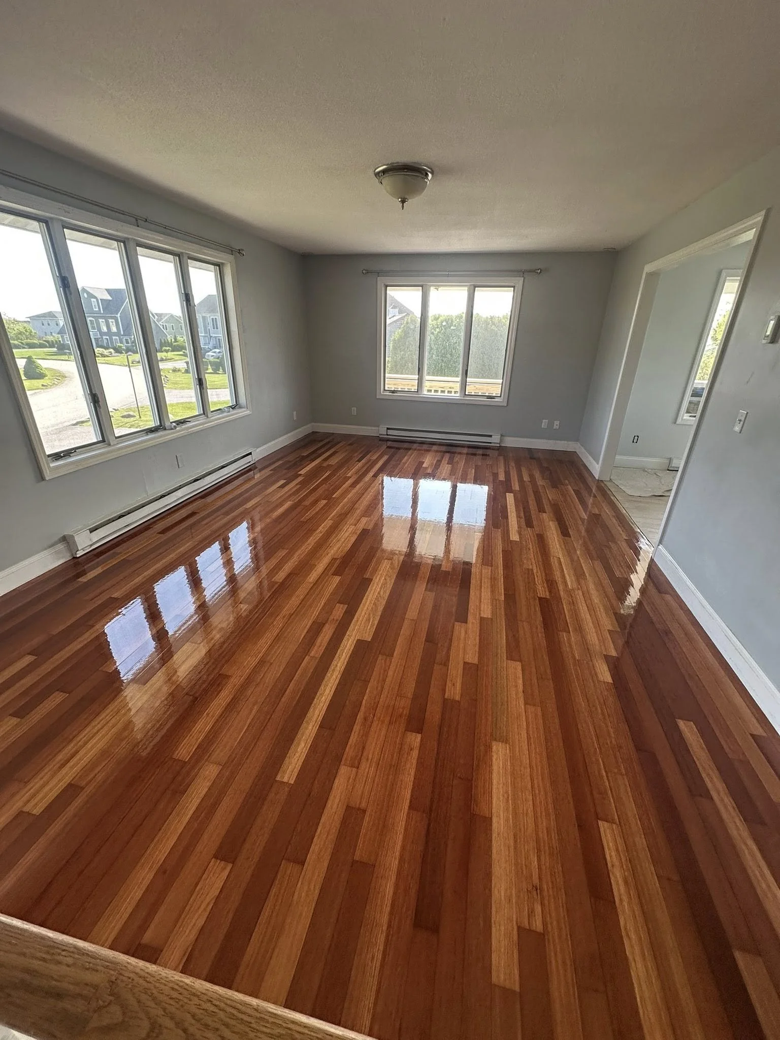 Empty living room with polished hardwood floors, white walls, large windows on two sides, a ceiling light fixture, and a view of a residential neighborhood outside.