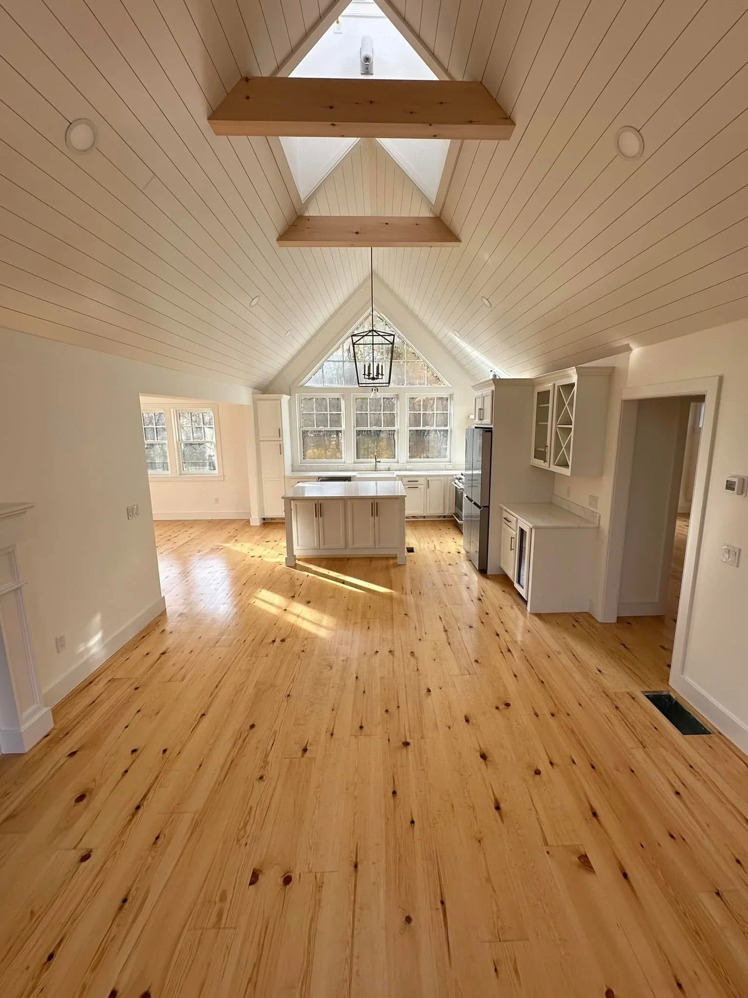 Spacious kitchen with vaulted ceiling, light wood flooring, white cabinets, a kitchen island, and large windows in the background.