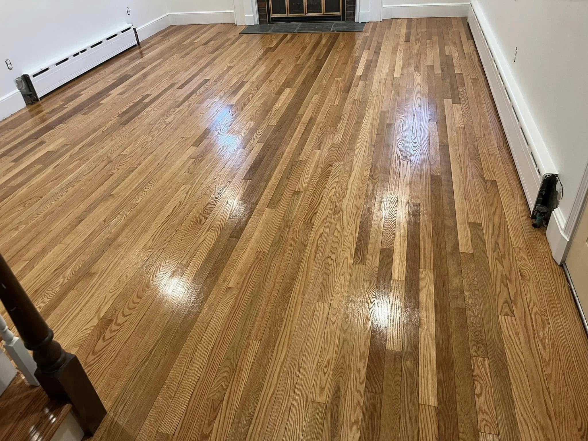 A hardwood floor with a natural finish in a room, with a white baseboard heater along the wall and a fireplace in the corner.
