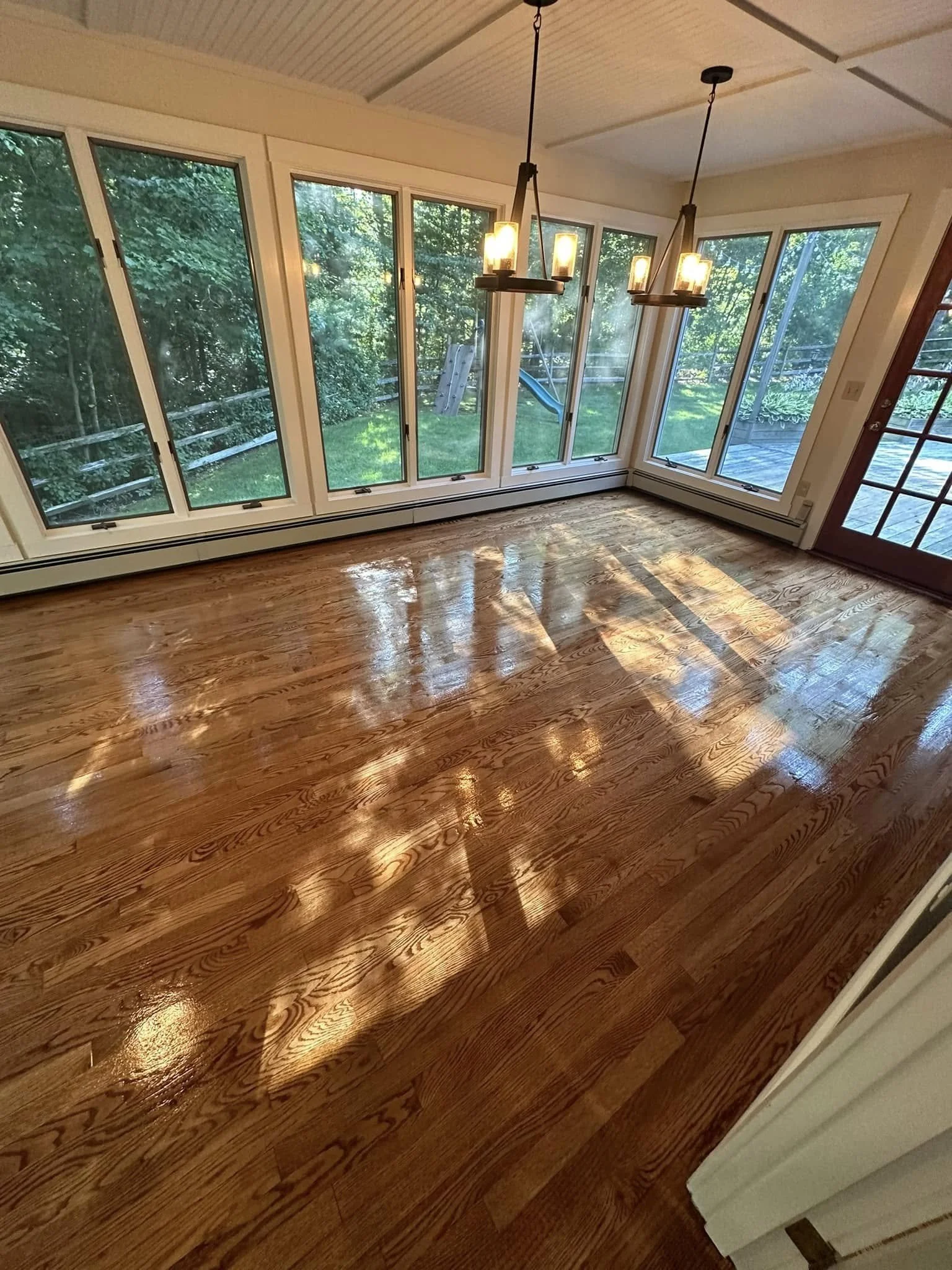 Empty room with polished wooden floor, large windows with greenery outside, and pendant lighting fixtures hanging from the ceiling.