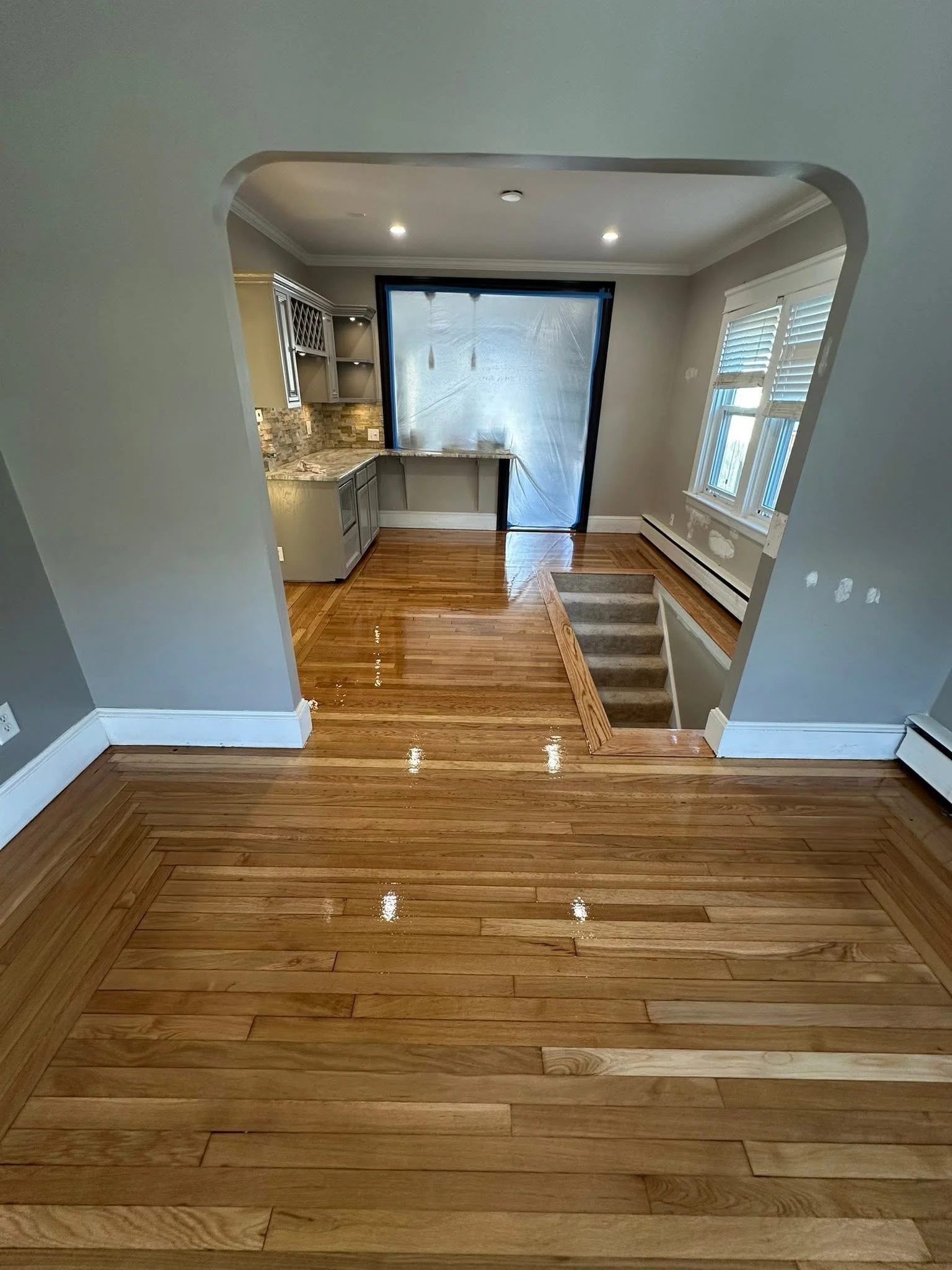 Empty room with hardwood floors, an open staircase, and a kitchen area with gray cabinets and a brick backsplash. A plastic sheet covers a window or doorway, and there are some windows with blinds on the right.