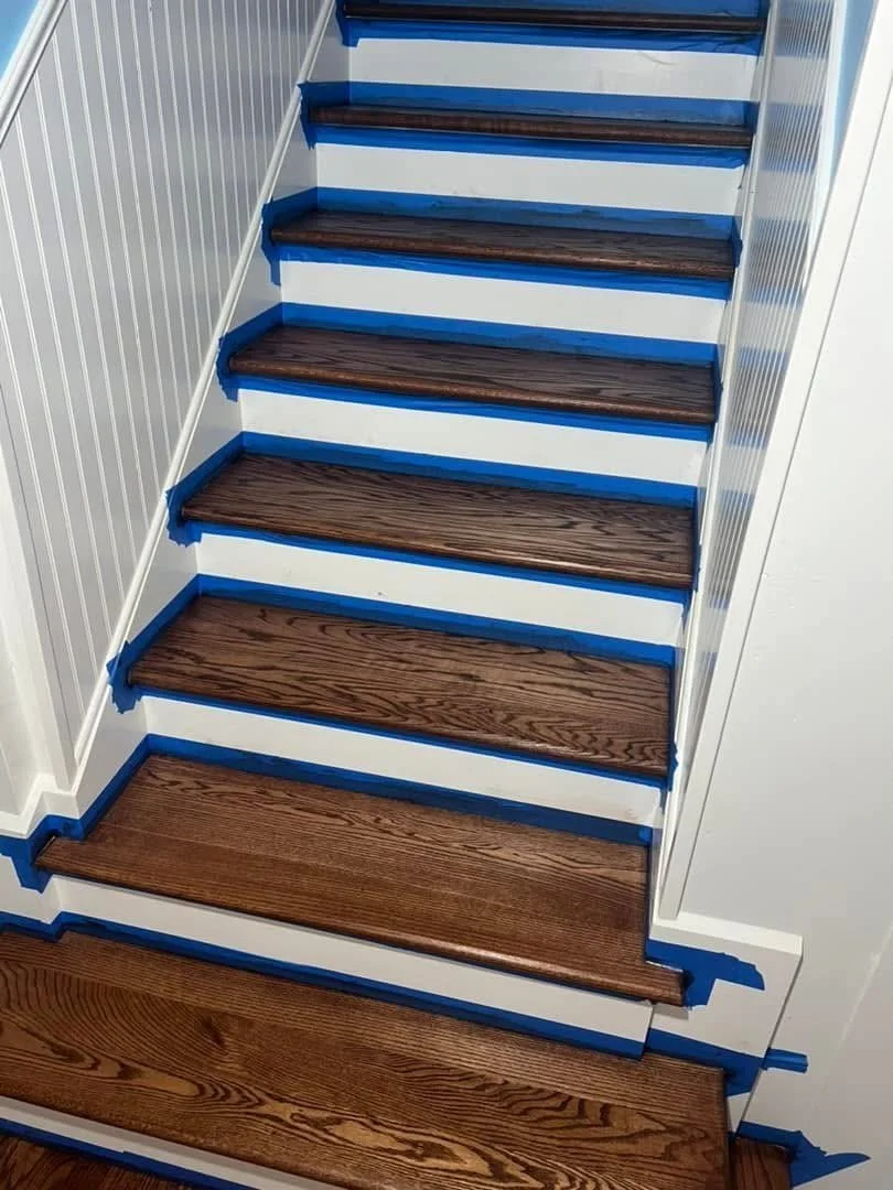Wooden staircase with dark brown steps, blue painter's tape along the edges, and white walls with vertical paneling.