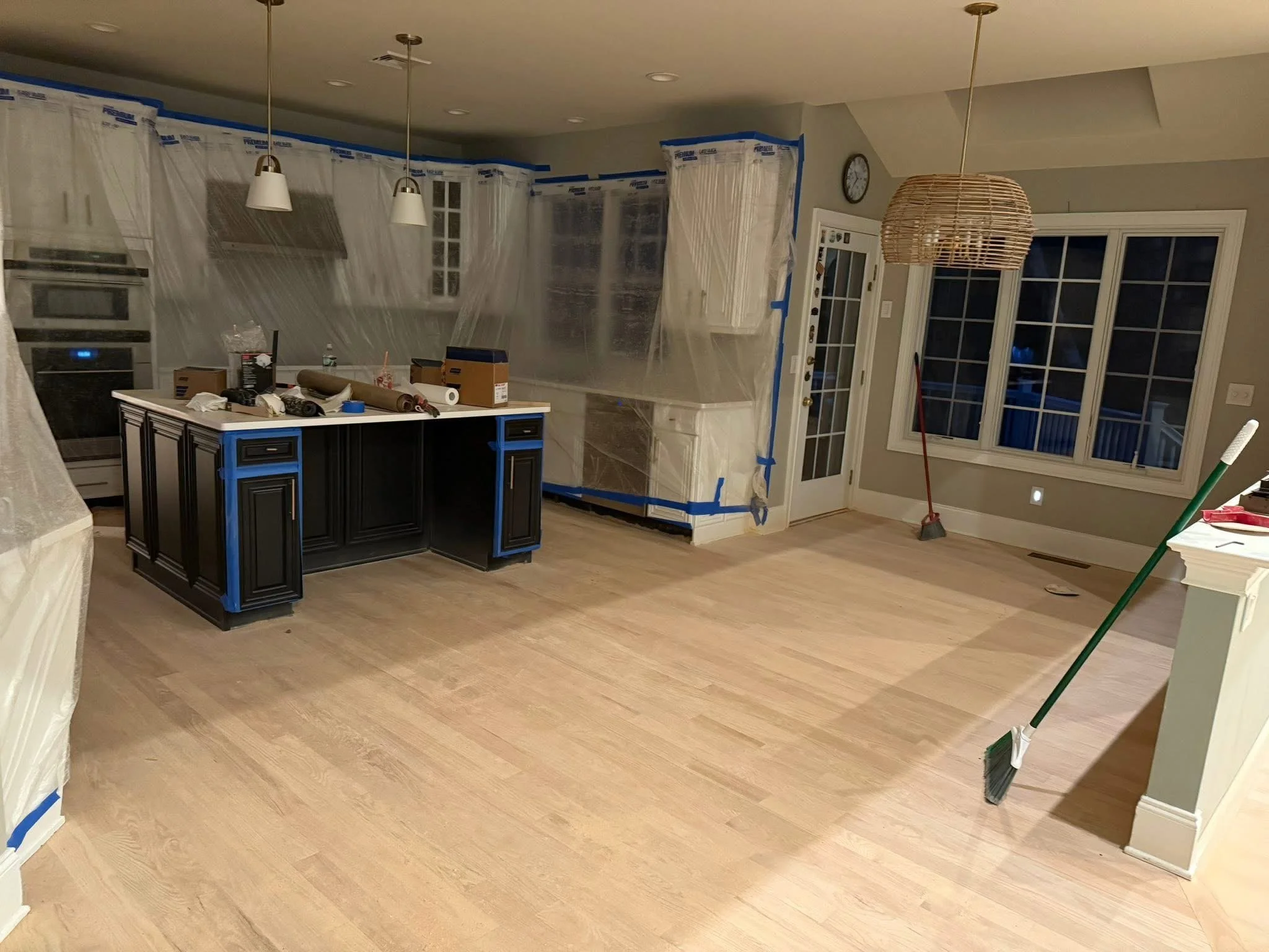 An indoor kitchen space undergoing renovation with plastic sheeting covering cabinets and part of the wall, a kitchen island, cleaning tools like a broom and mop, and various boxes and supplies on the island and countertops.