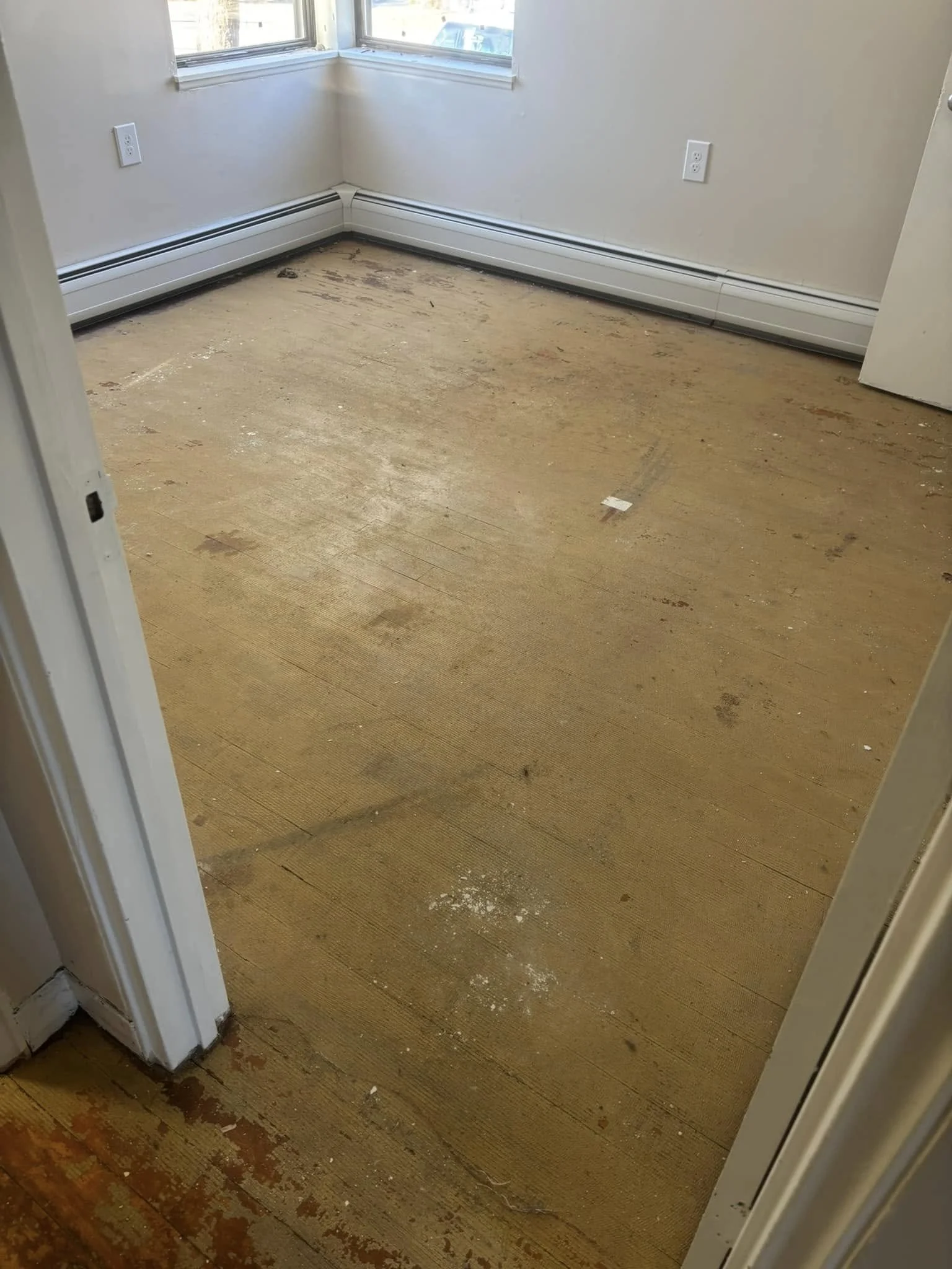 Empty room with hardwood floor, two windows, and white baseboard heater, wall outlets visible.