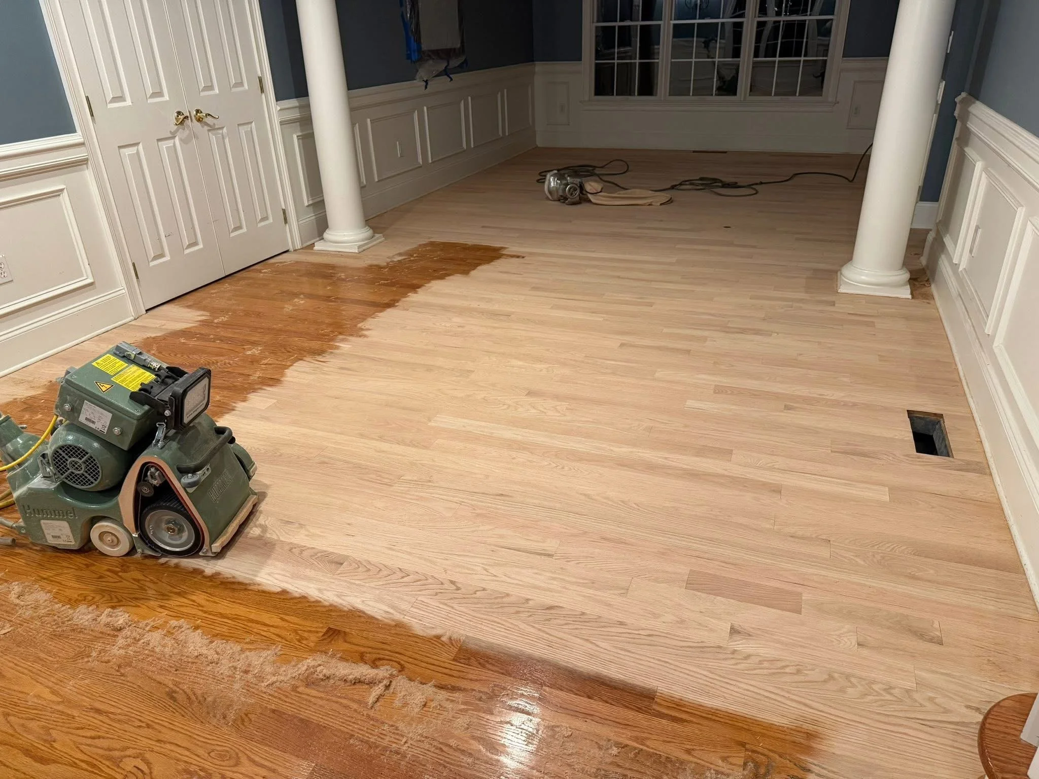 A room with newly installed light wood flooring, some of which is still being sanded or refinished, with a floor sander machine and other tools present.