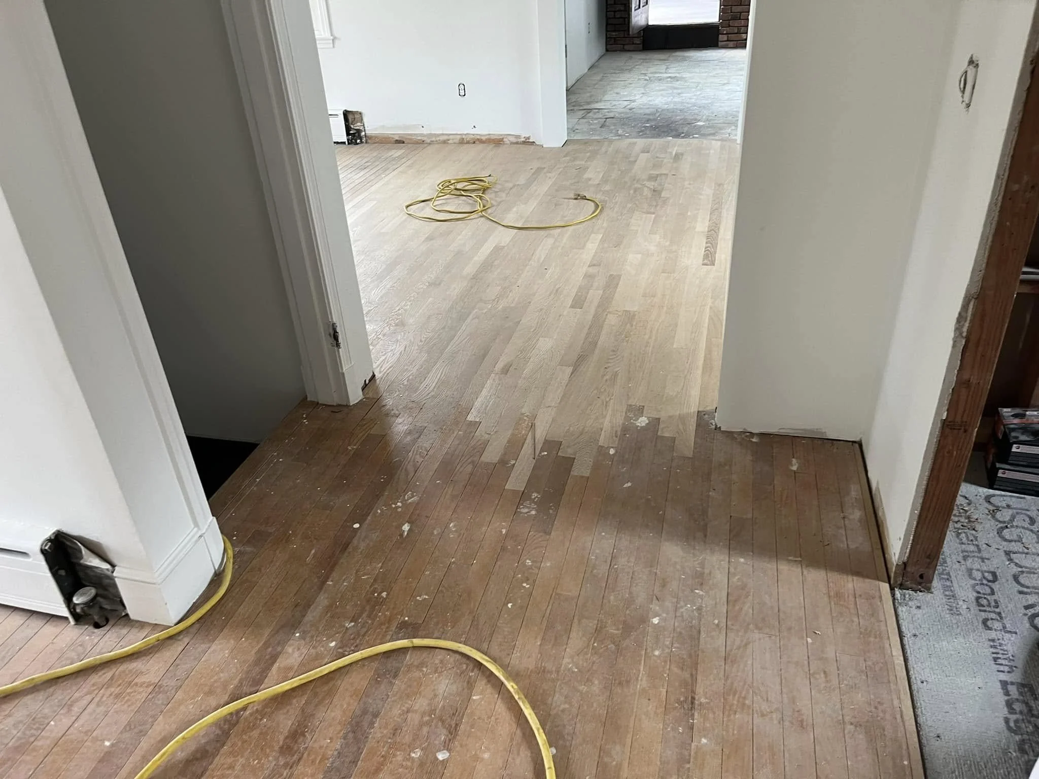 Interior view of a room under renovation with wooden flooring; some sections are unfinished; yellow extension cords on the floor.