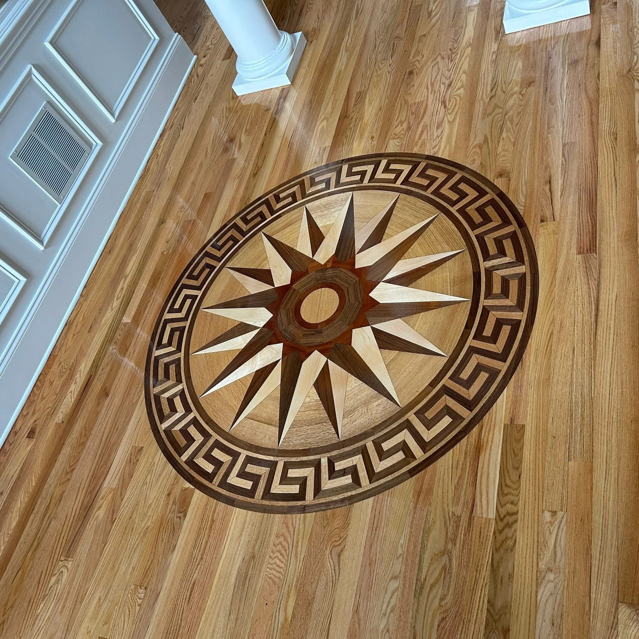 Decorative wooden floor medallion with a starburst pattern and Greek key border, on hardwood floor.