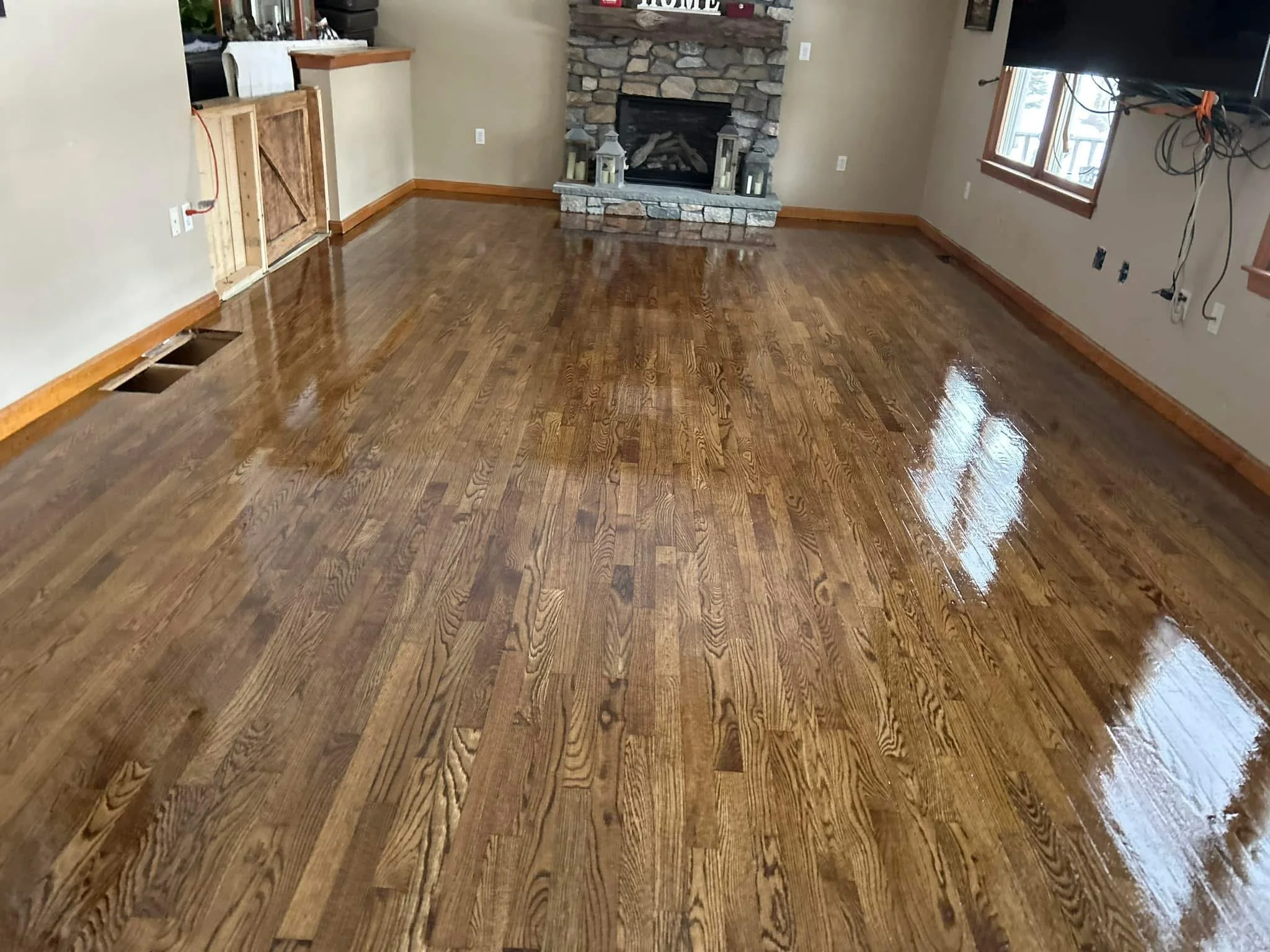 Freshly polished wooden floor in a living room with a stone fireplace, a window, and some furniture in the background.