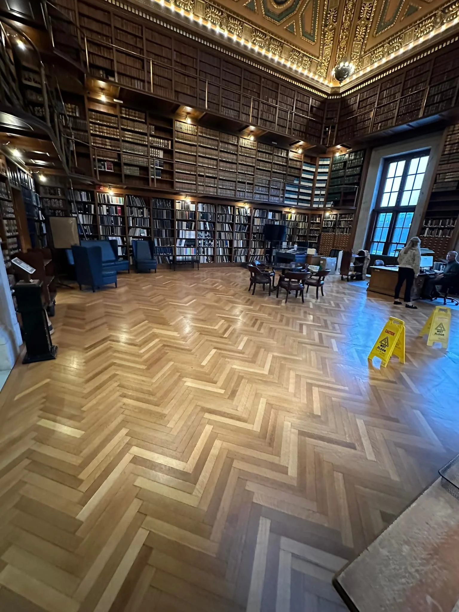 A large, elegant library room with tall wooden bookshelves filled with books covering the walls from floor to ceiling. The room has a parquet wood floor, a few chair seating areas, a computer desk with chairs, and large windows allowing natural light. There are also some people and yellow caution wet floor signs.