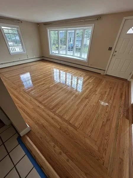 Empty living room with polished hardwood floor, large window with view of outside, and a white front door.