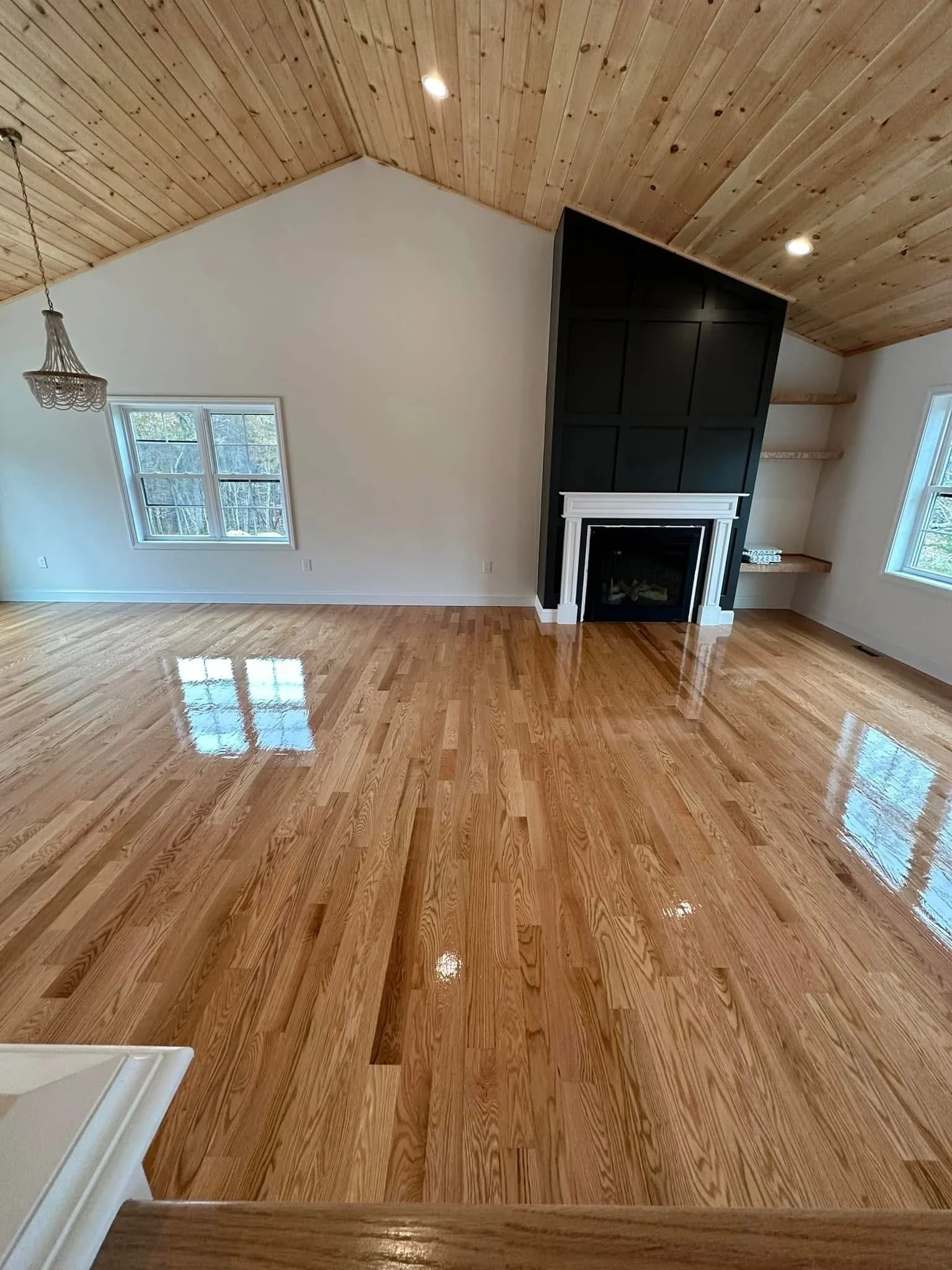 Empty living room with polished hardwood floors, white walls, a wood-paneled vaulted ceiling, windows, and a black accent wall with a fireplace and built-in shelves.