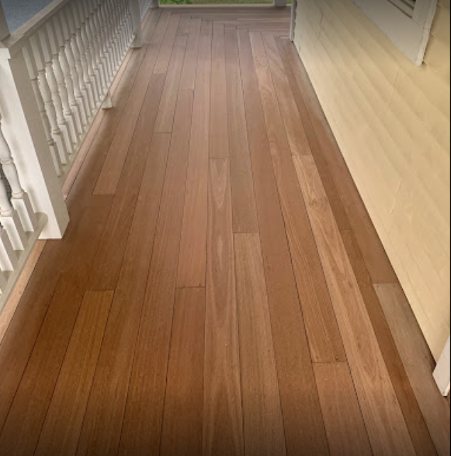 Close-up view of a polished hardwood floor on a porch or balcony with a railing on the left and a yellow wall on the right.