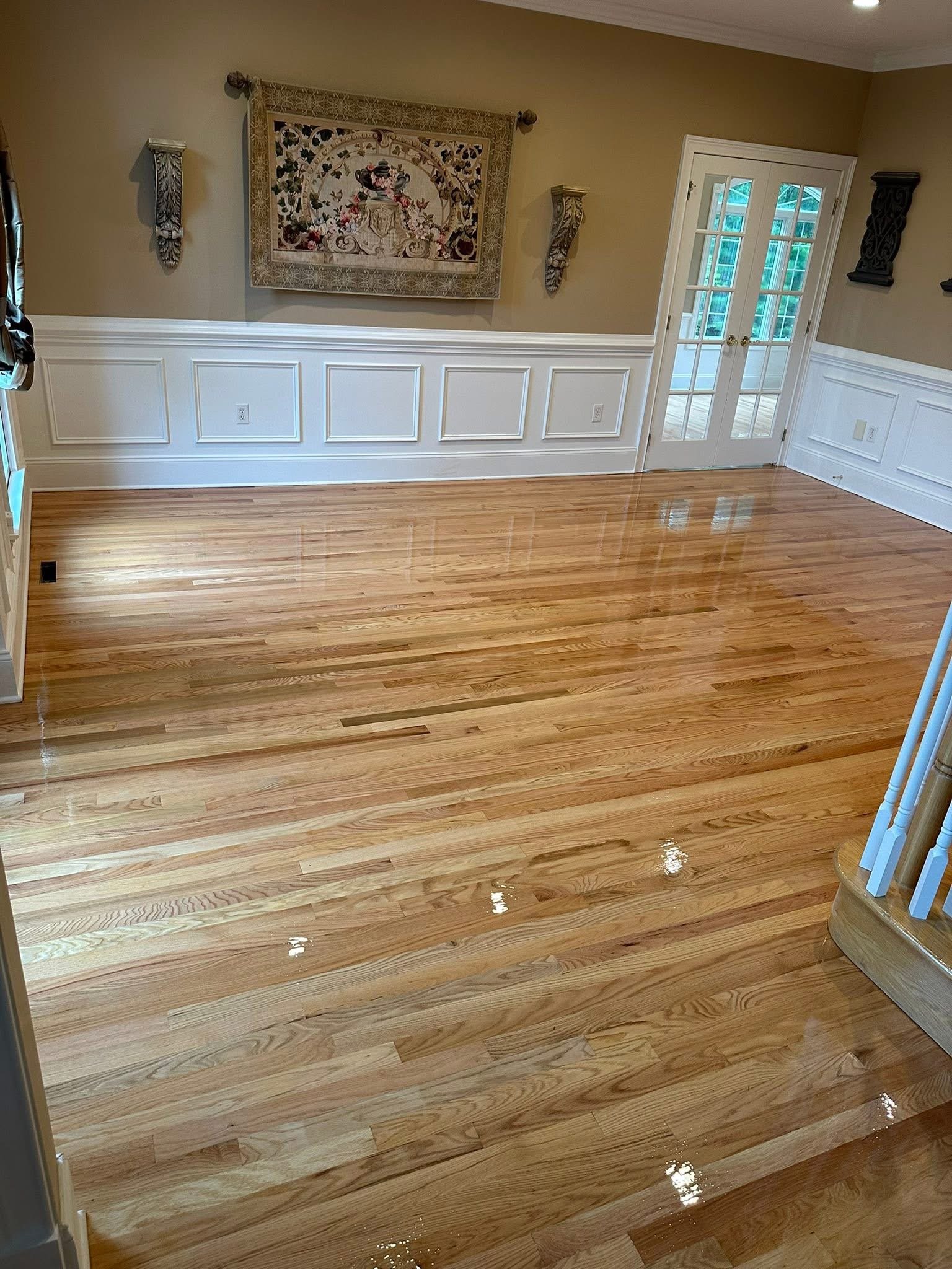 A freshly polished hardwood floor in a dining room with beige walls, white wainscoting, and a tapestry hanging on the wall. There are double French doors leading to an outdoor area in the background.