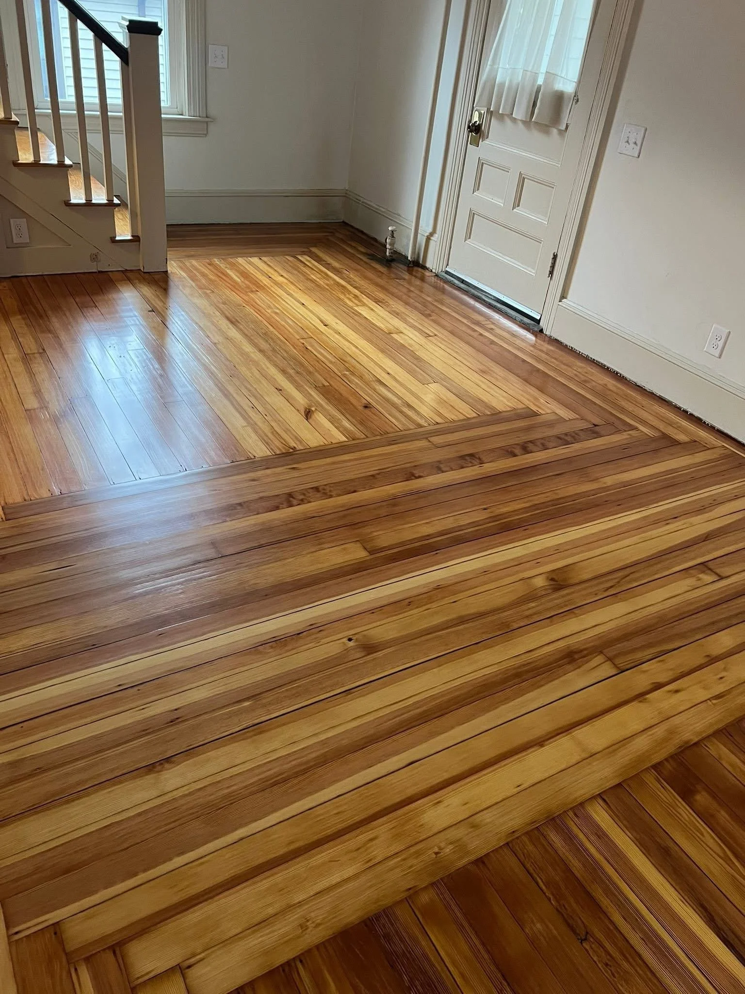 Room with hardwood flooring under renovation, part of the floor raised with new planks, white walls, a door with a curtain, a window, and stairs in the corner.