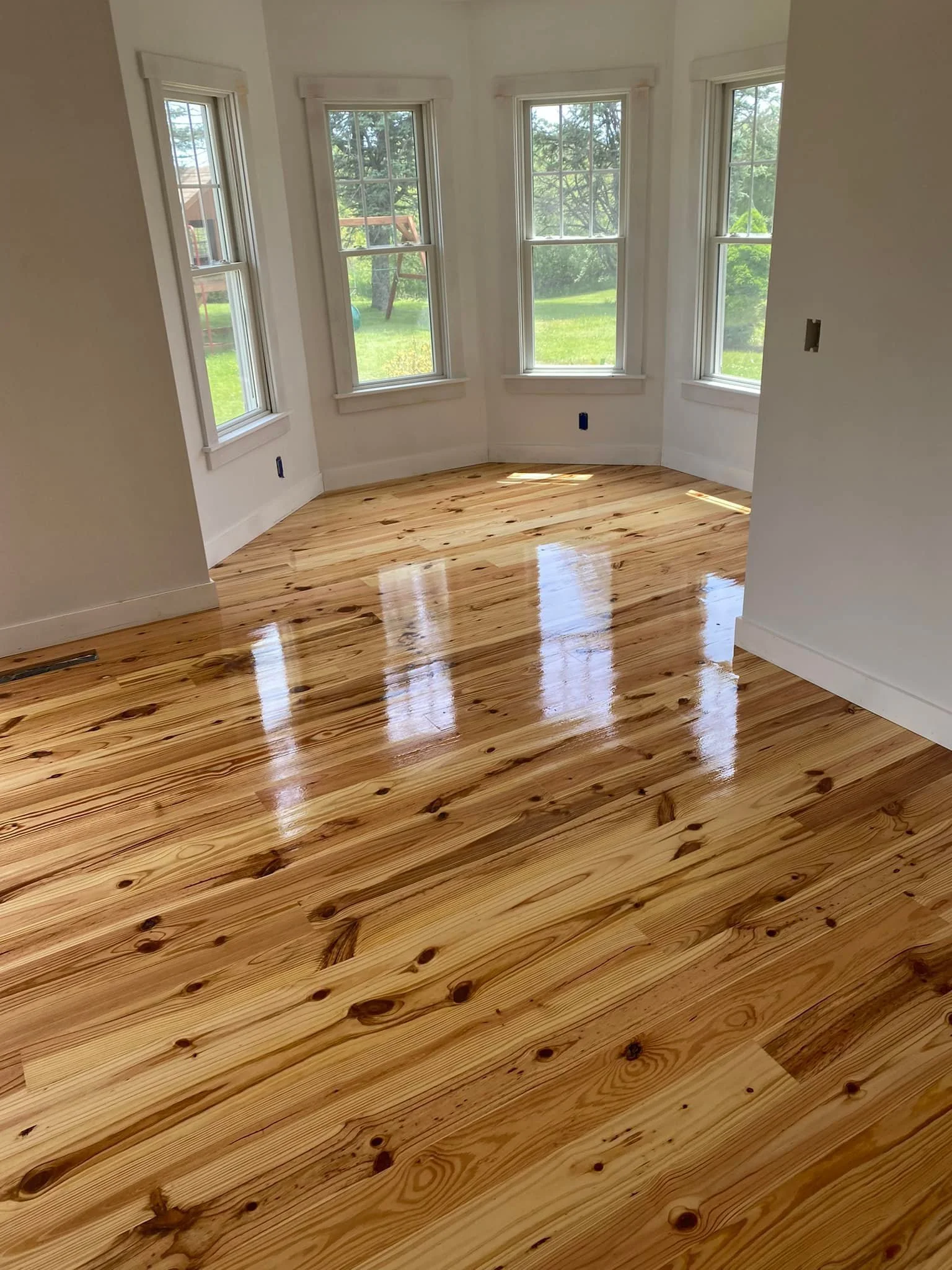 A room with three large windows and a shiny hardwood floor, with a view of a green backyard outside.