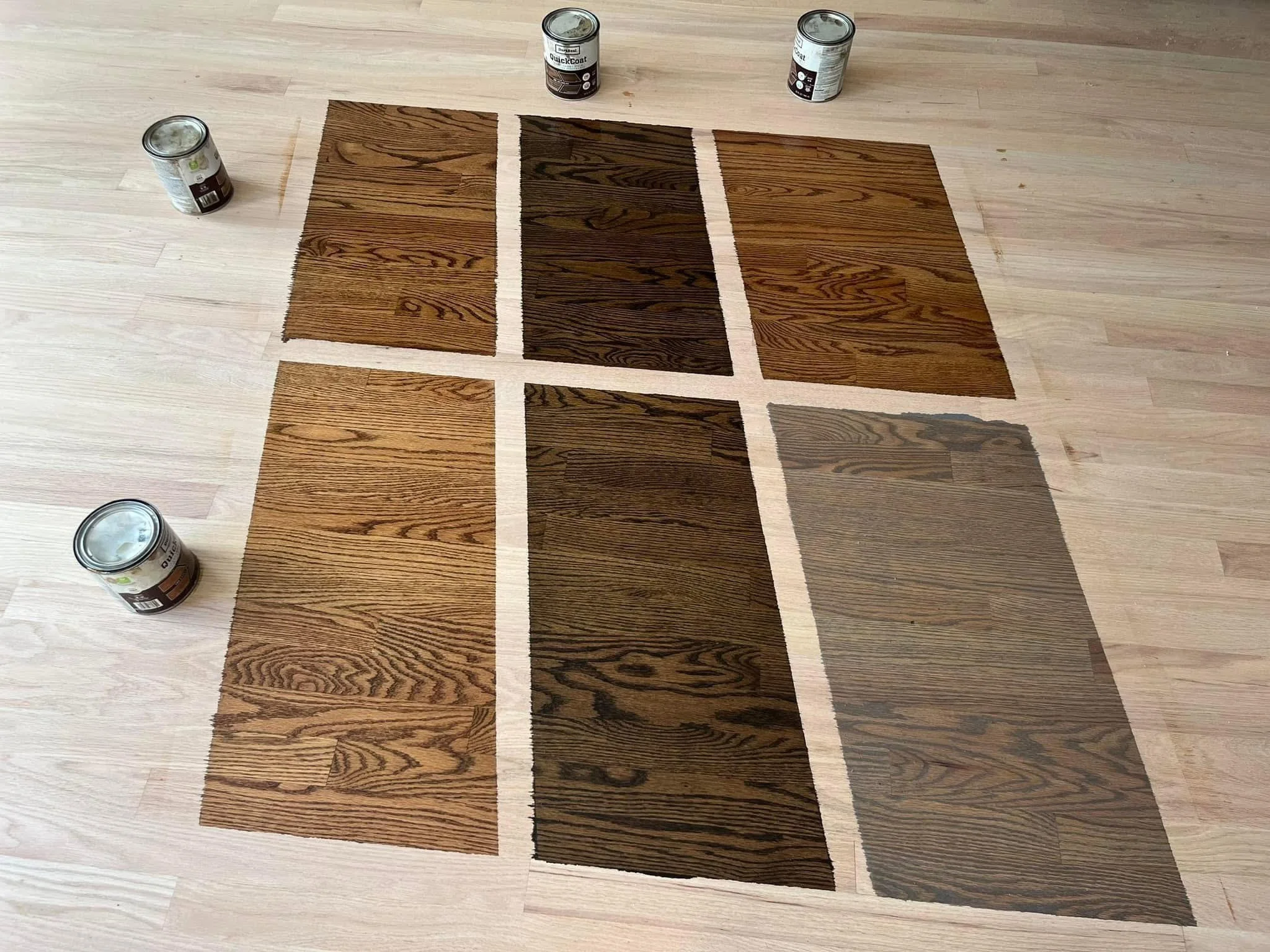Six pieces of wood flooring samples with varying shades of brown, arranged on a light wood floor. Four cans of wood stain or finish are positioned around the samples.