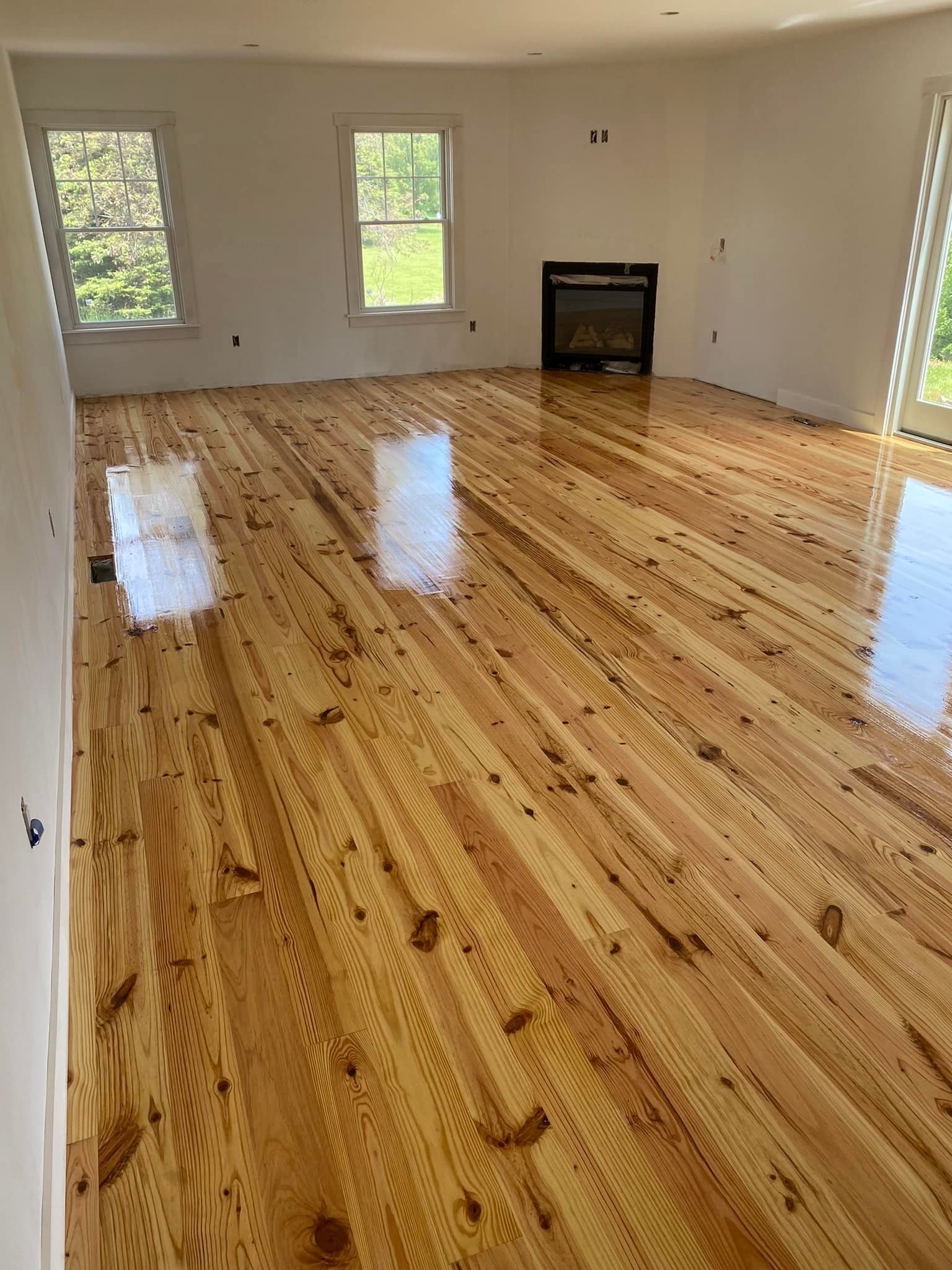 Empty living room with newly installed shiny hardwood floors, white walls, three windows, and a fireplace.