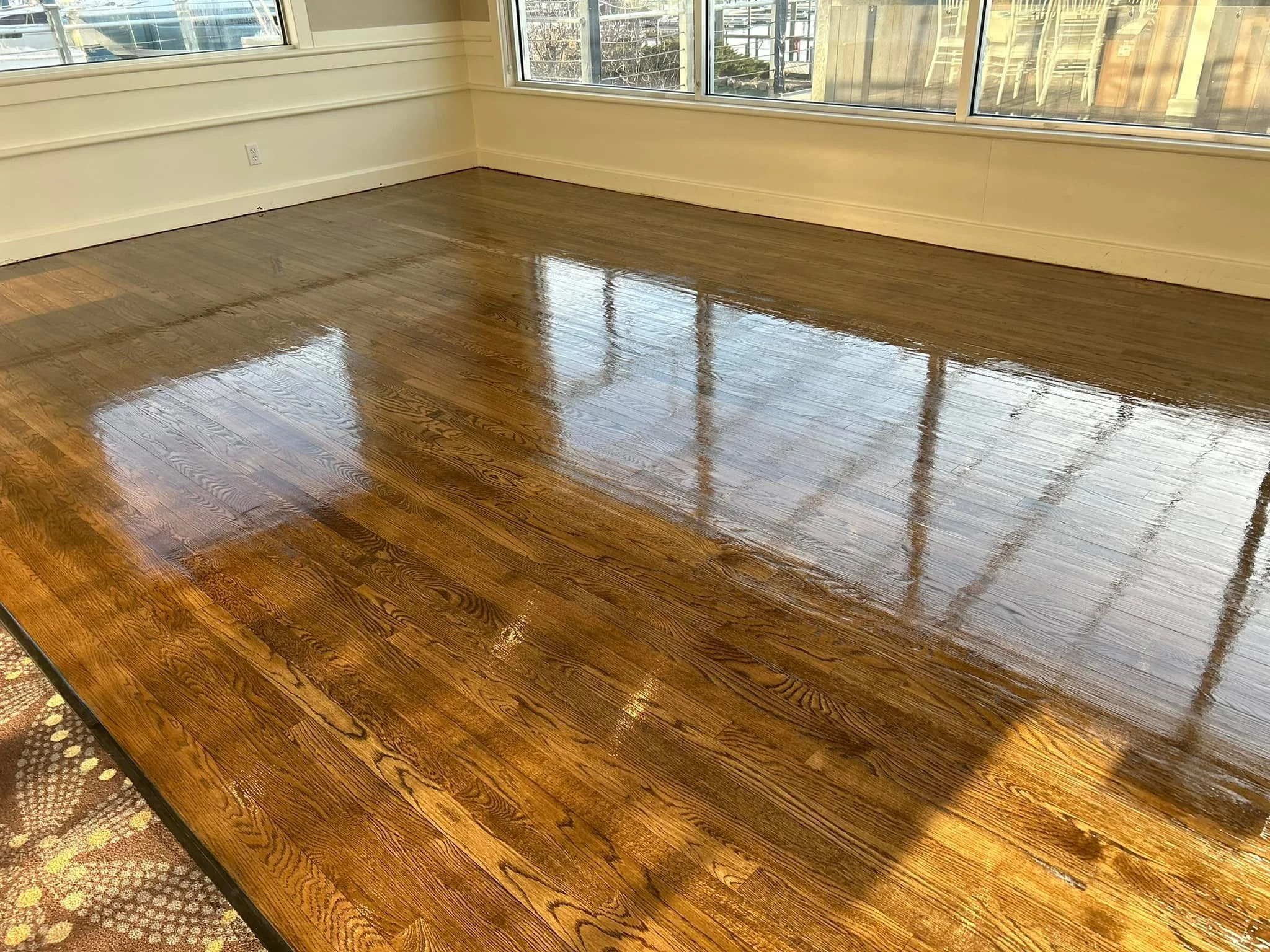 A room with polished hardwood floors, white walls, large windows with sunlight, and a view of a balcony or outdoor area.